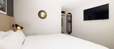 Una habitación de hotel con una cama, televisión montada en la pared, lámpara de noche y un espejo.