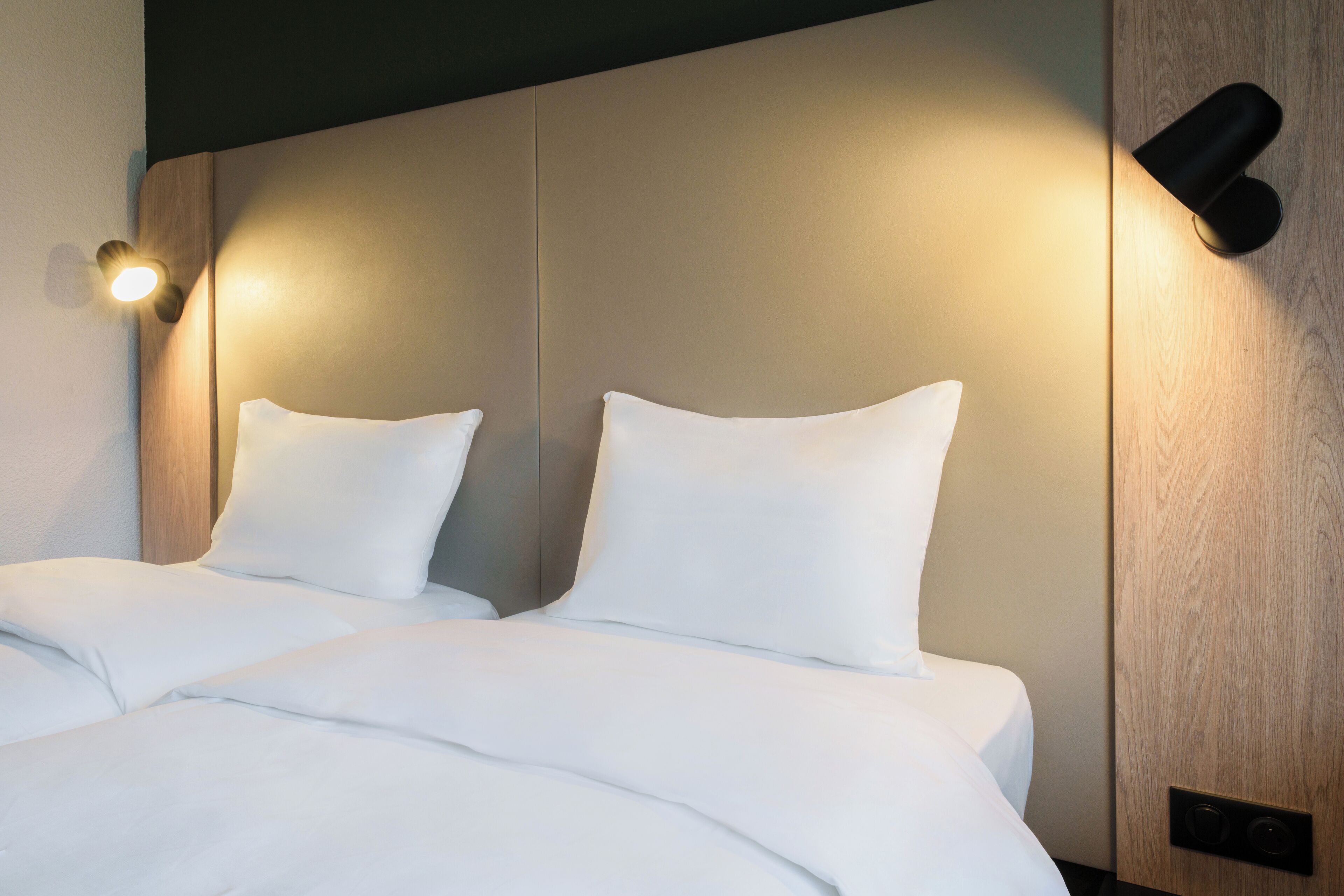 A neatly made bed with white linens and two pillows, a wall-mounted light fixture, and a wooden panel on the wall.