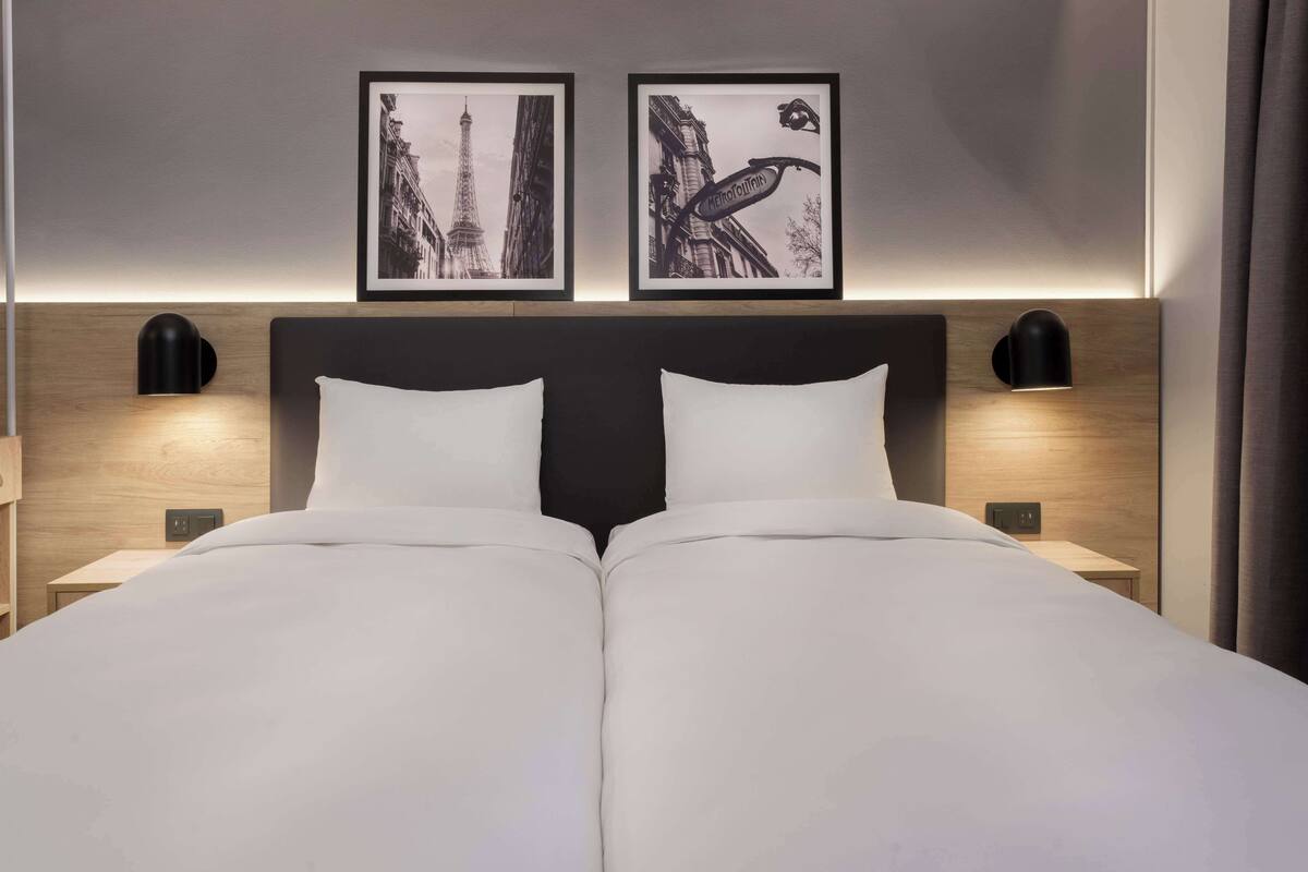 Een tweepersoonsbed met witte lakens, twee zwarte wandlampen en twee ingelijste foto's van Parijse architectuur.