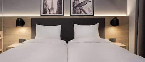 A double bed with white linens, two black wall-mounted lamps, and two framed pictures of Parisian architecture.