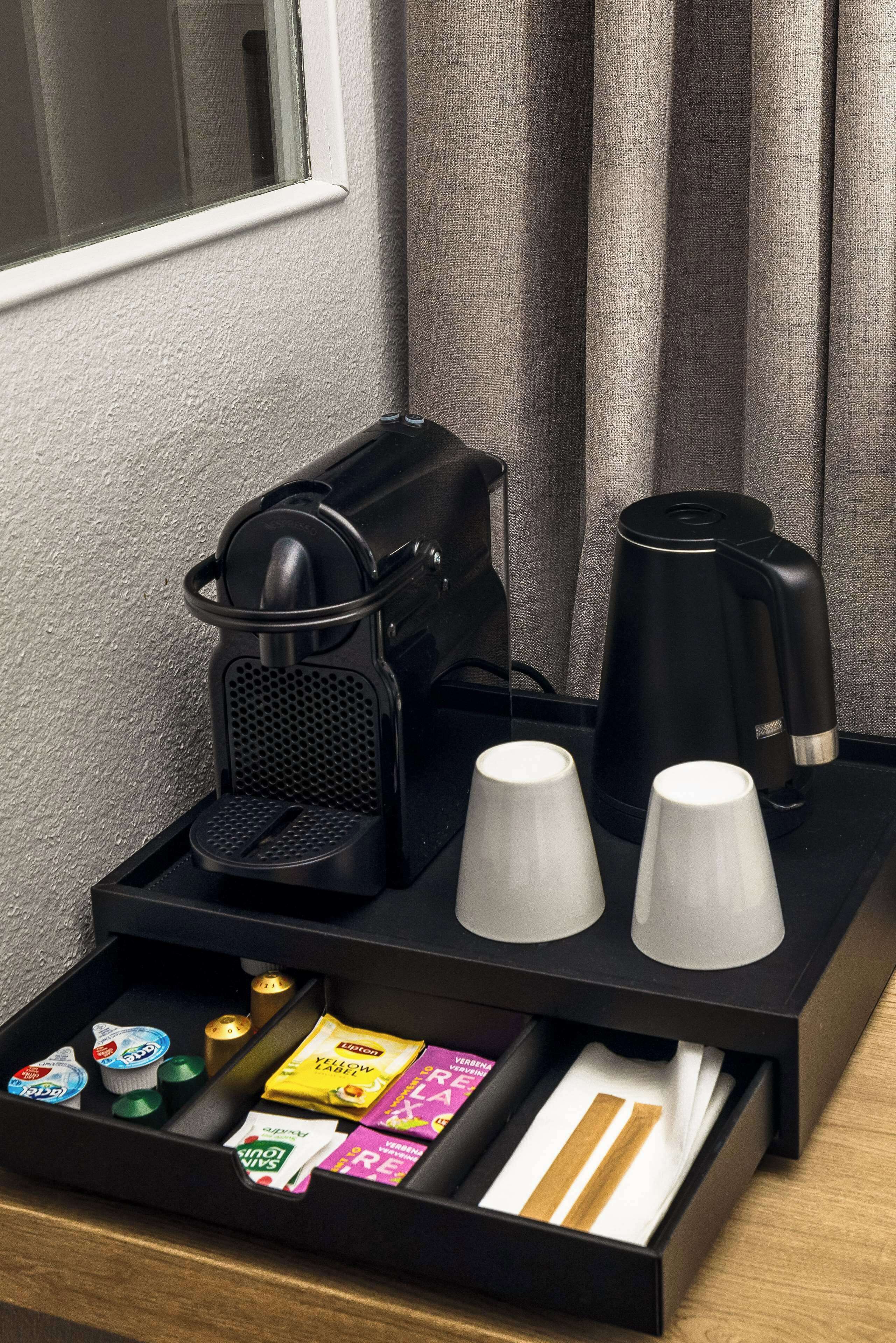 Un mini-bar dans une chambre d’hôtel, avec une machine à café, deux tasses blanches et divers en-cas et boissons.
