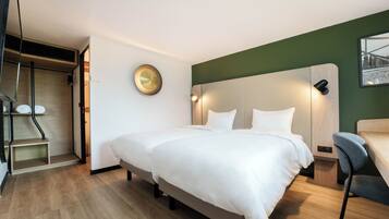 A modern hotel room with a large bed, a desk, and a chair. There is a small shelf and a picture on the wall.