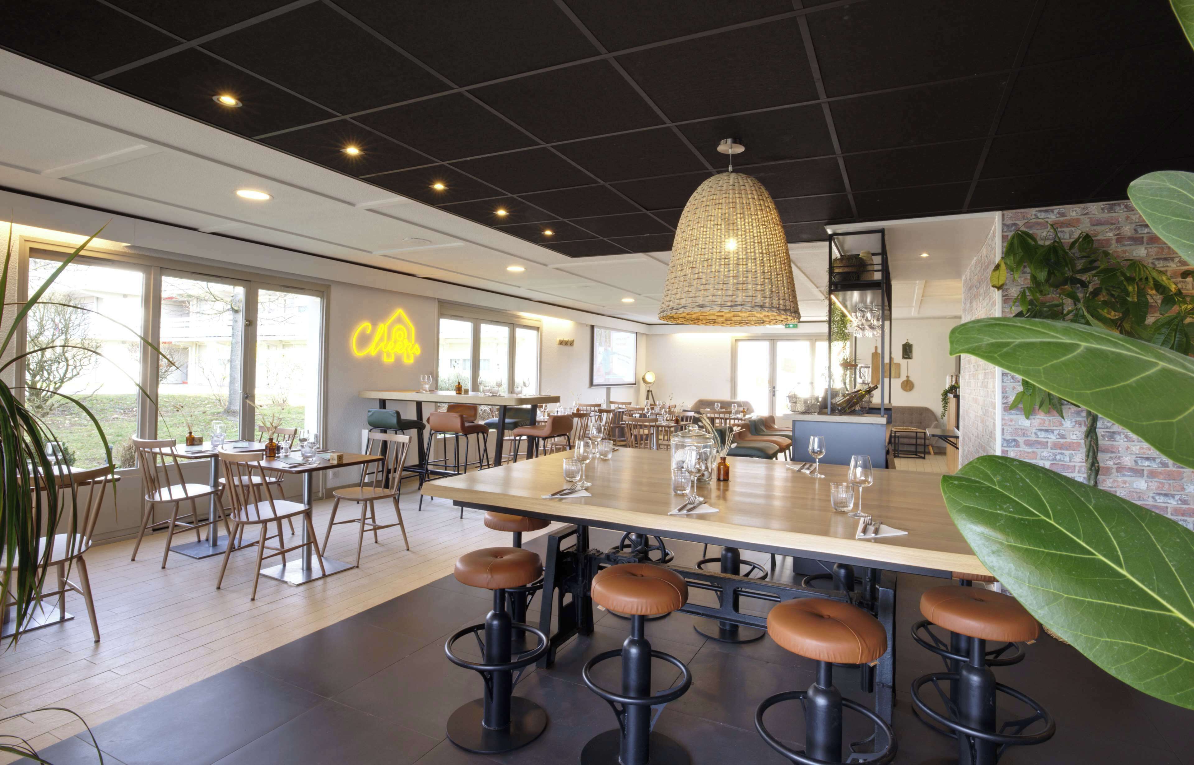 A modern dining area with a large table, multiple chairs, and bar stools. There are large windows providing natural light and a view of the outdoors.