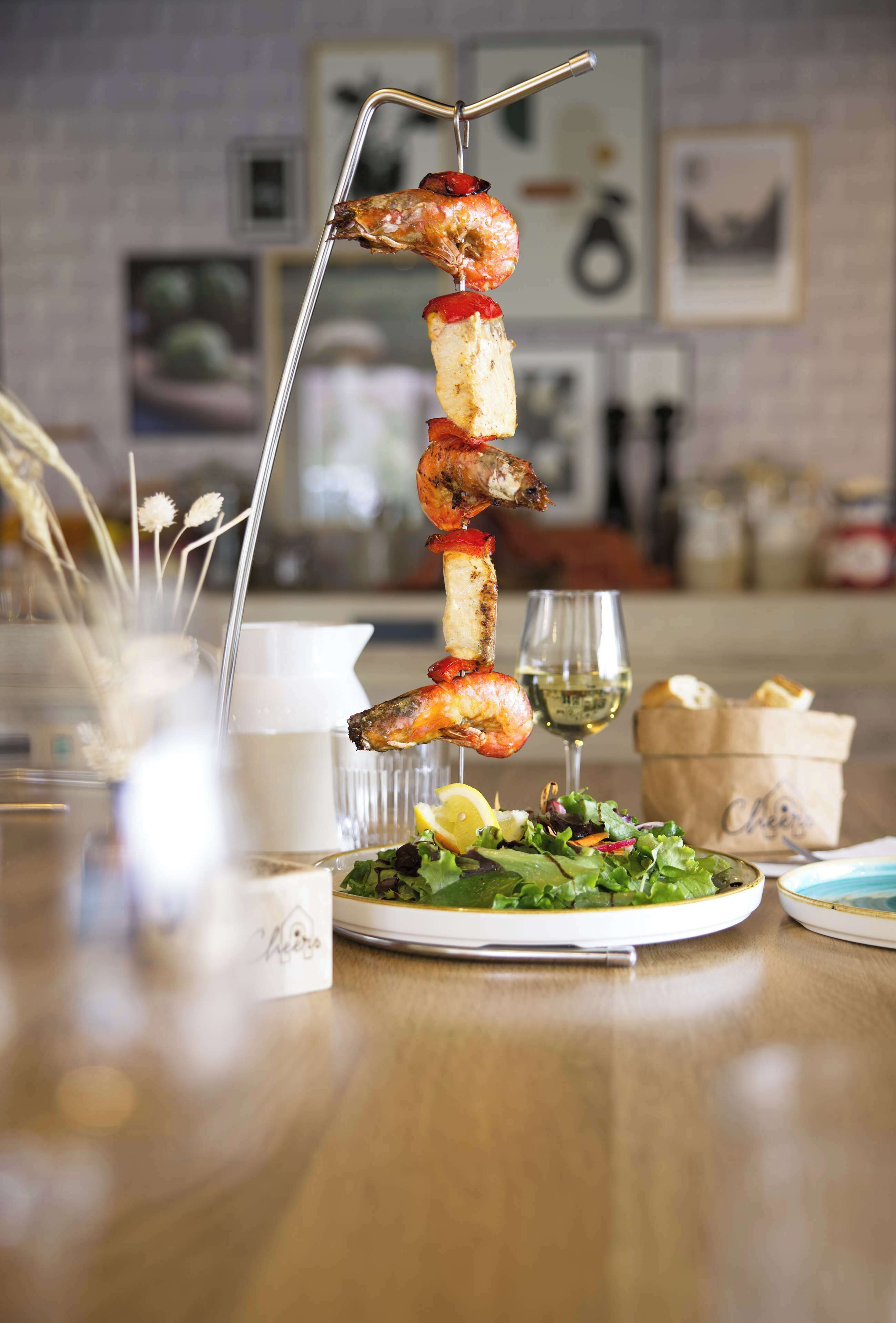 Une brochette de nourriture présentée sur un présentoir, un verre de vin blanc et une assiette de salade sur une table en bois.