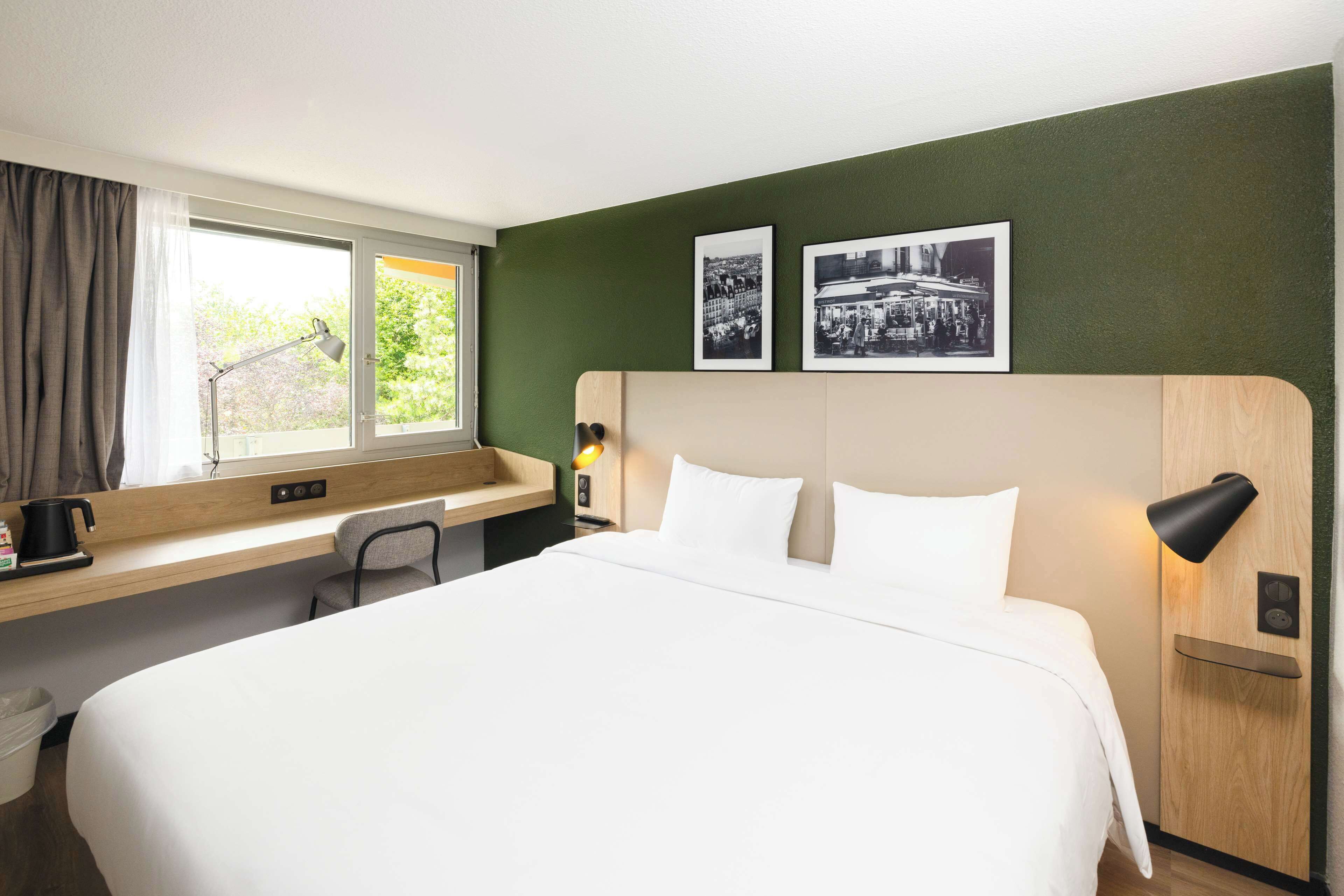 A hotel room with a large bed, a desk with a chair, a window with curtains, and two framed pictures on the wall.