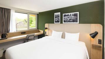 A hotel room with a large bed, a desk with a chair, a window with curtains, and two framed pictures on the wall.