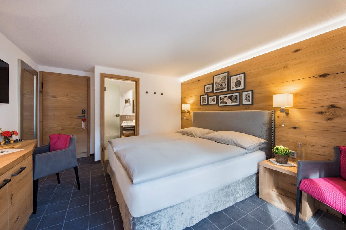A modern bedroom with a large bed, a desk, and a chair. There are framed pictures on the wall and a small table with a plant.