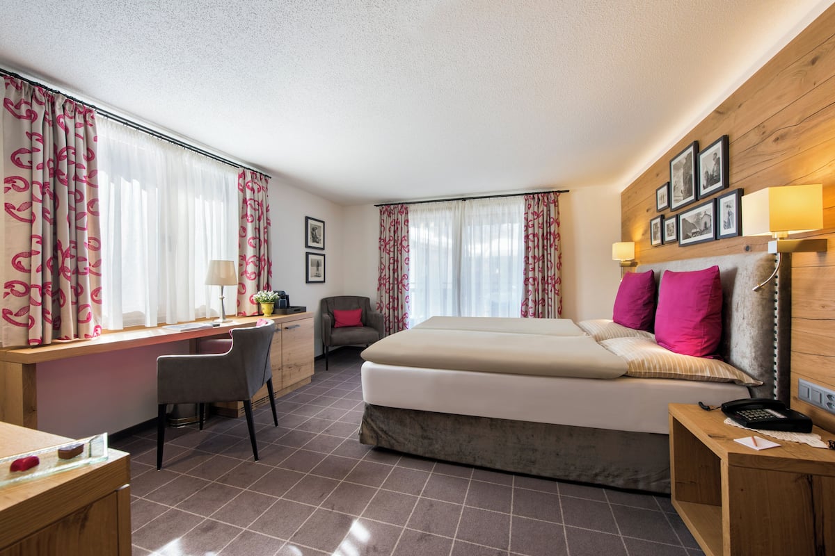 A hotel room with a bed, desk, chair, and a small table with a lamp.