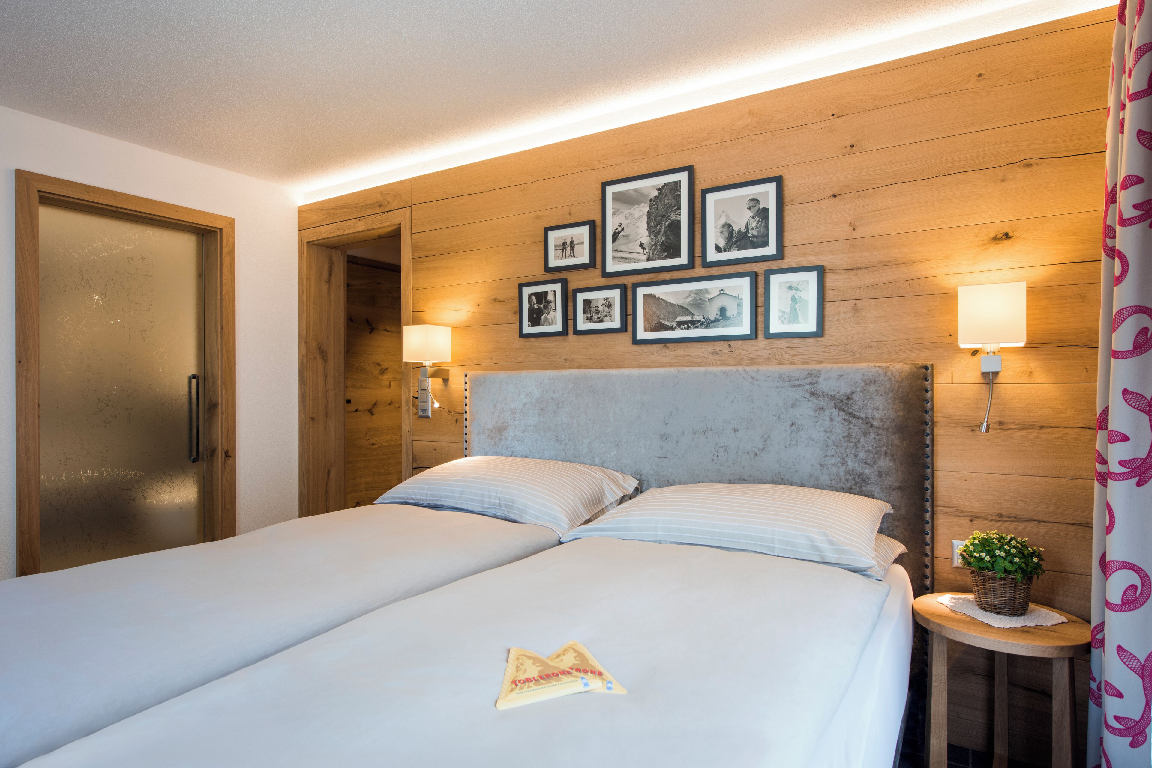 Hotellihuone, jossa on parivuode, puinen pääty, seinällä kehystettyjä tauluja, kaksi yöpöytävalaisinta ja pieni pöytä, jonka päällä on kasvi.