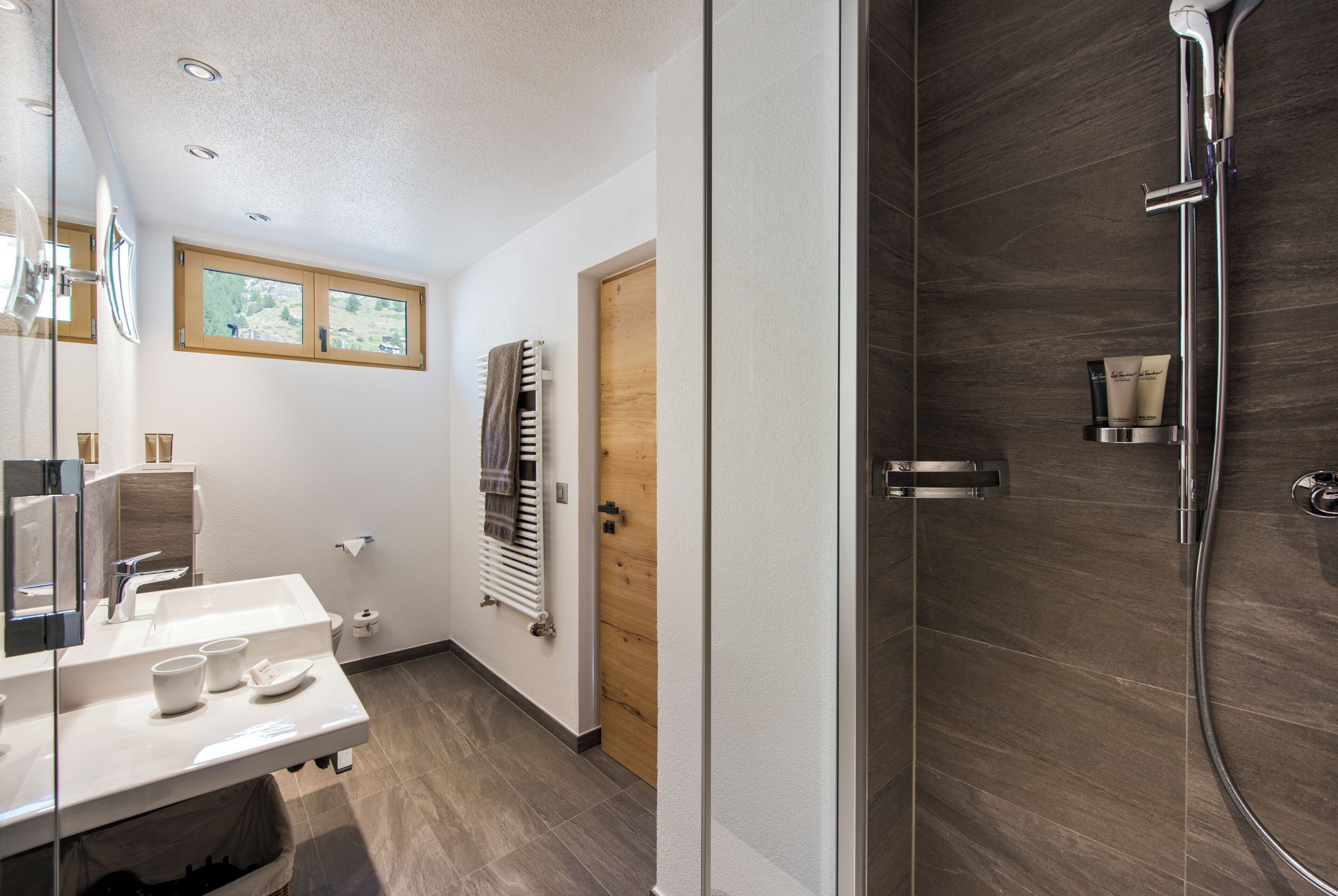 Une salle de bain moderne dotée d’une douche à parois de verre, d’une porte en bois et d’une fenêtre laissant entrer la lumière naturelle.