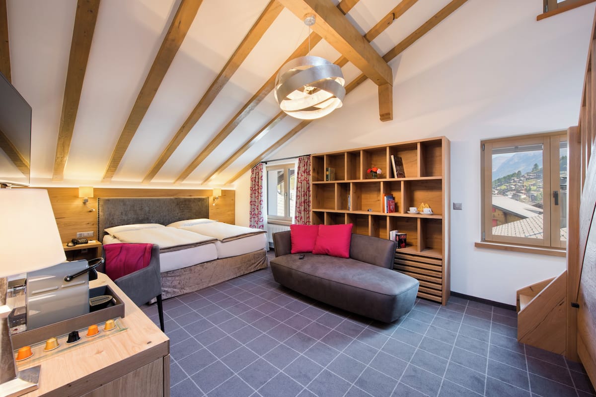 A cozy room with a bed, a sofa, a small table with a toaster, a bookshelf, and a window with a mountain view.