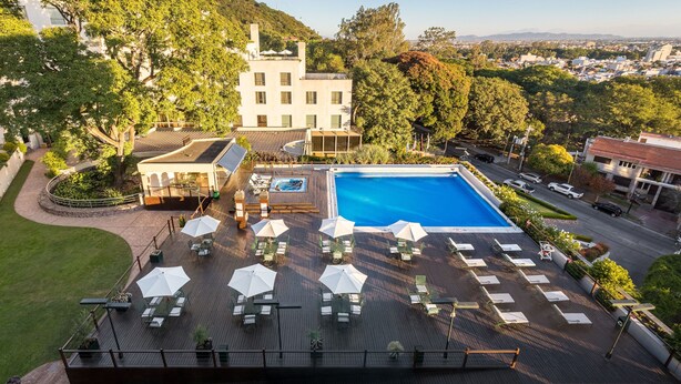 Un área de piscina en la azotea con sillones de descanso y sombrillas, rodeada de exuberante vegetación y vistas a la ciudad.
