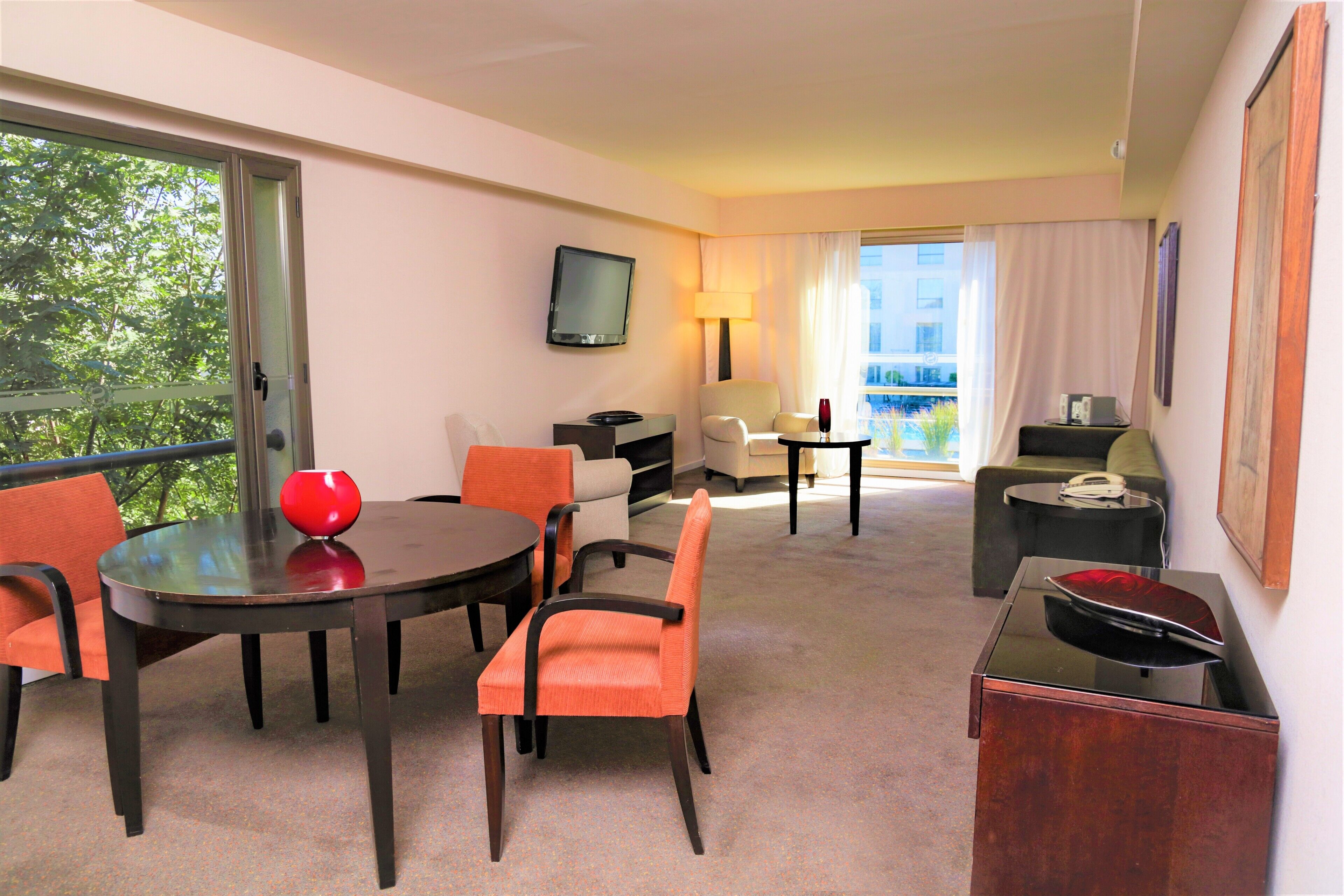 A hotel room with a dining area, a TV mounted on the wall, a sofa, a small table with a lamp, and a view of the outdoors through the window.