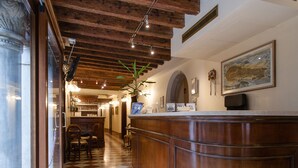 Eine Hotelhalle mit Schachbrettmuster-Boden, Holzbalken an der Decke, einer Rezeption und einem Essbereich im Hintergrund.