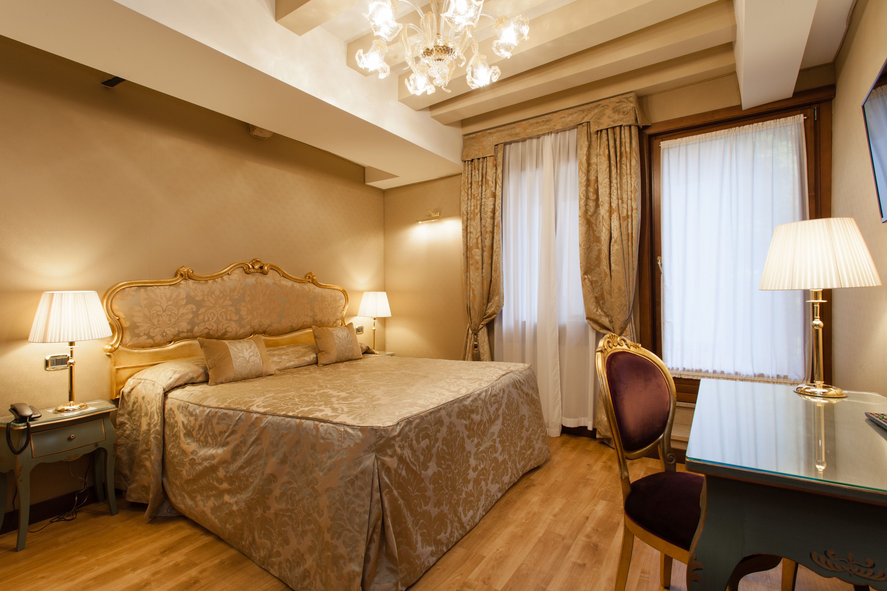 A hotel room with a large bed, two bedside tables with lamps, a desk with a chair, and a chandelier.