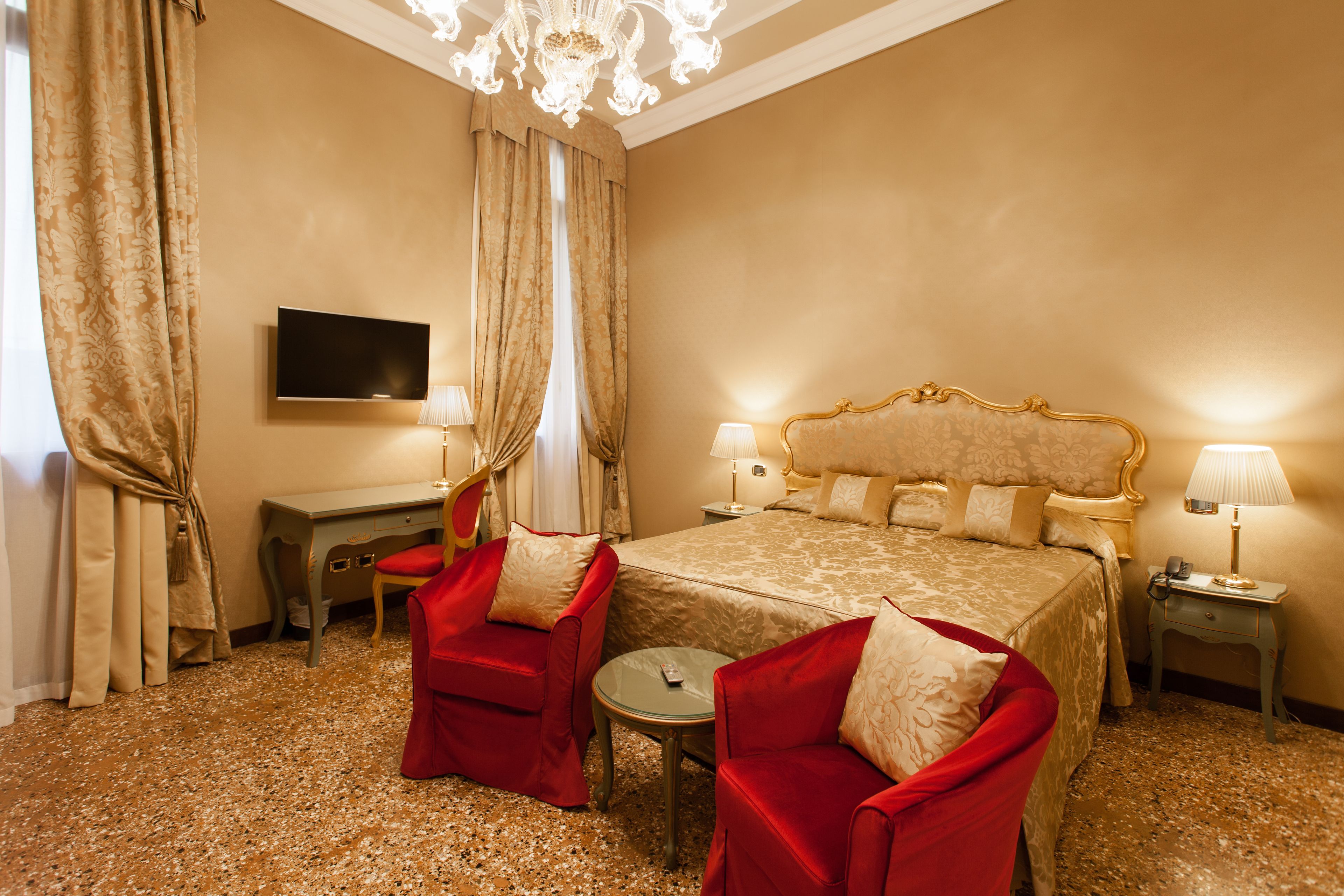 A hotel room with a large bed, two red armchairs, a small round table, a desk with a flat-screen TV, and two bedside lamps.
