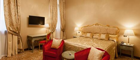 A hotel room with a large bed, two red armchairs, a small round table, a desk with a flat-screen TV, and two bedside lamps.