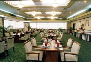 A hotel dining room with multiple tables and chairs, large windows, and a green carpet.