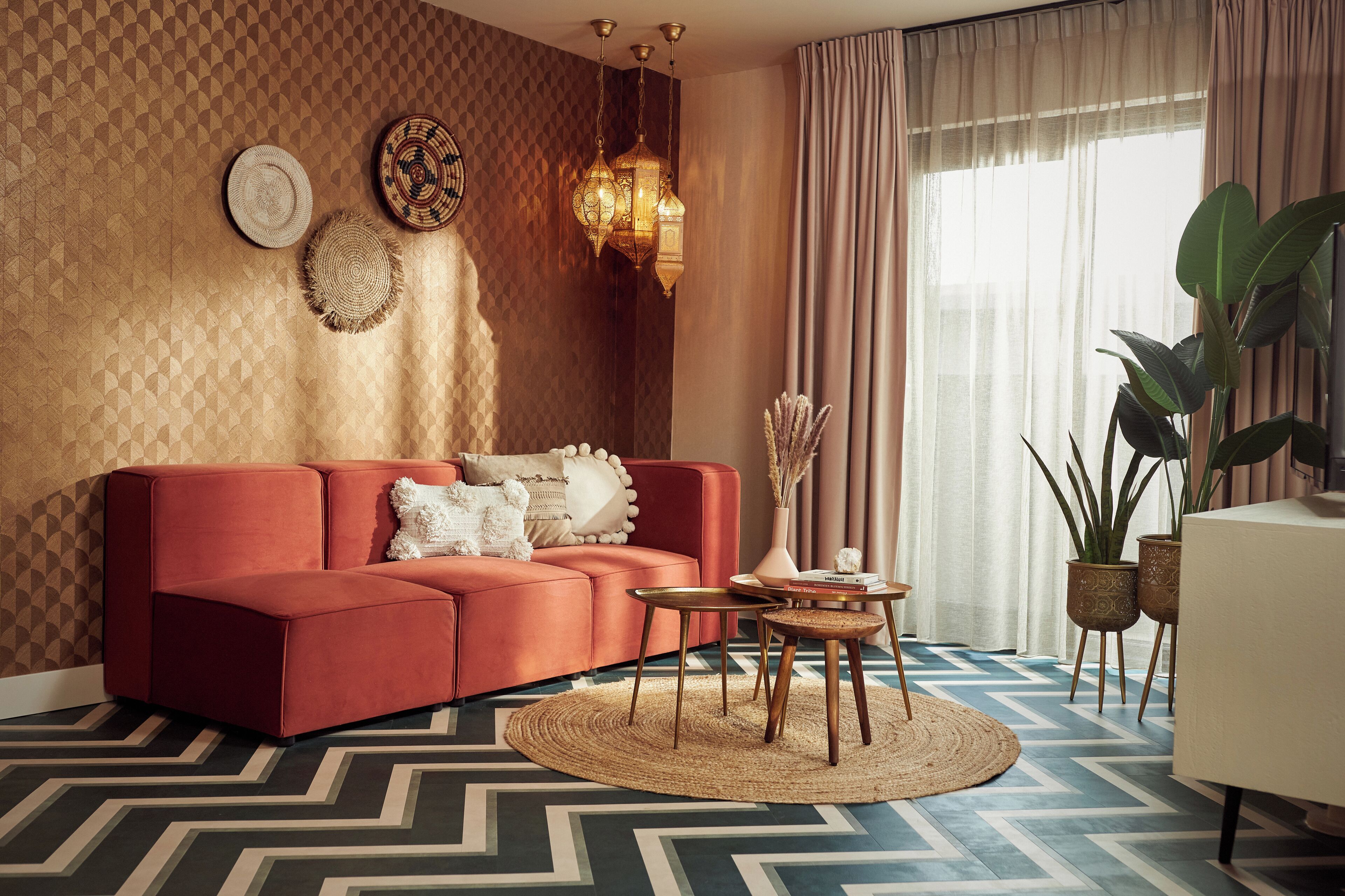 A modern living room with a red sofa, patterned wallpaper, a chandelier, and a large window with curtains.