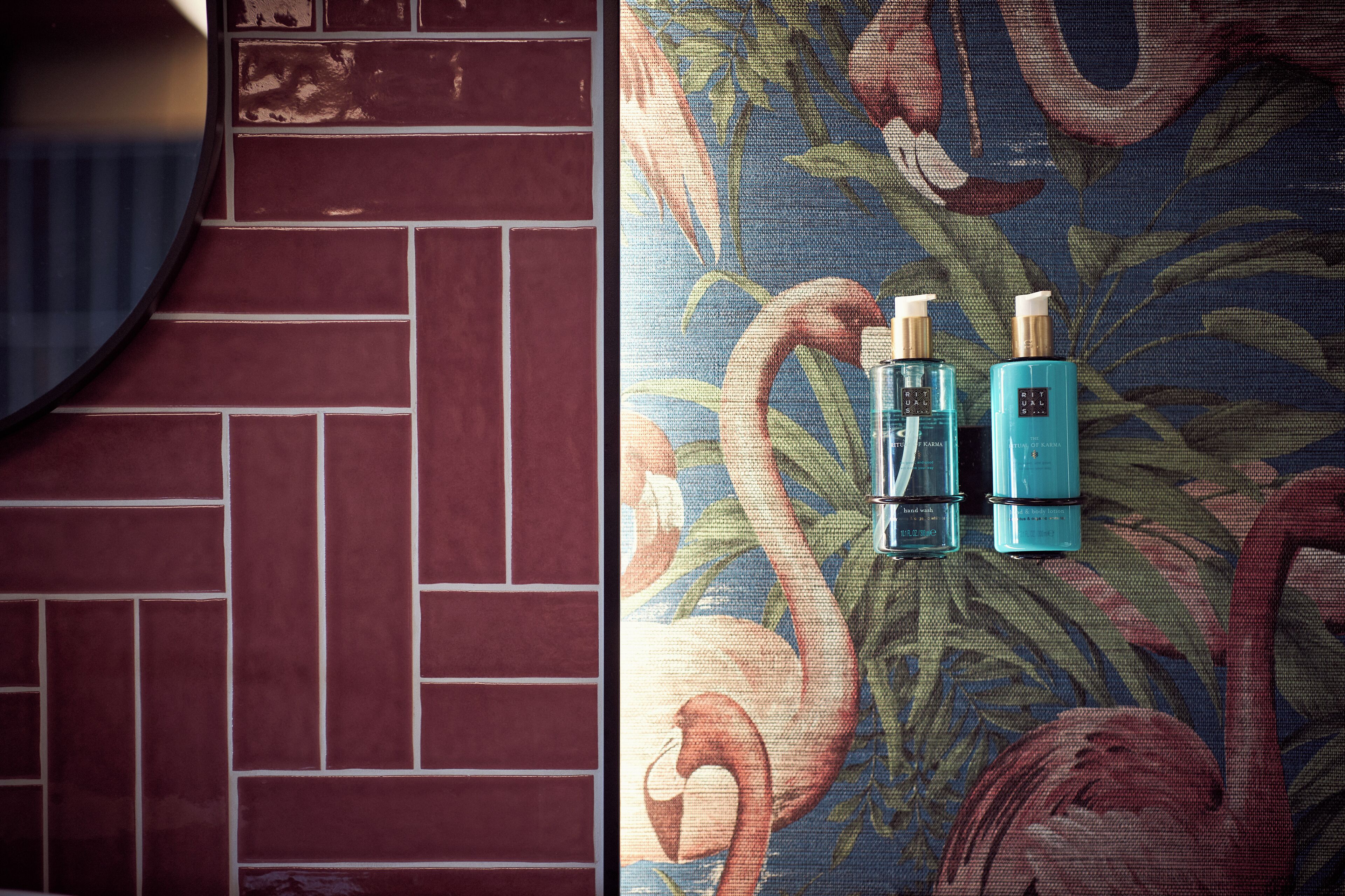A bathroom with a red tiled wall and a floral patterned wall. There are two blue bottles on the floral wall.