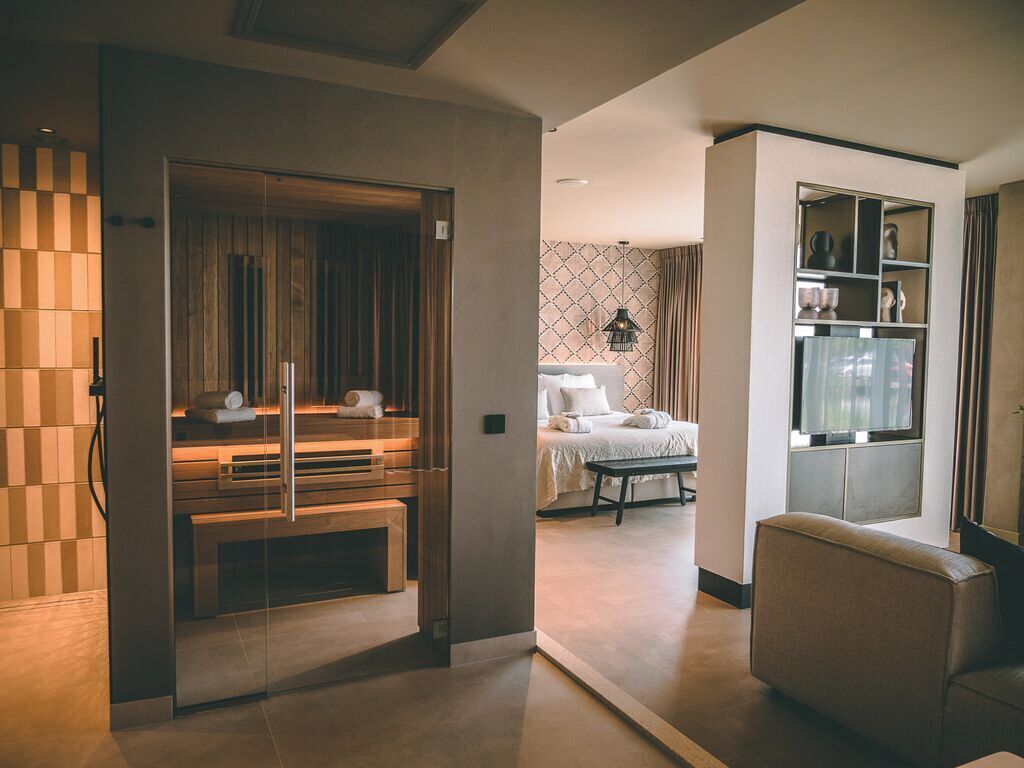 Una habitación de hotel moderna con un banco de madera, una cama con cabecera estampada, una mesita y un rincón de estar con sofá.