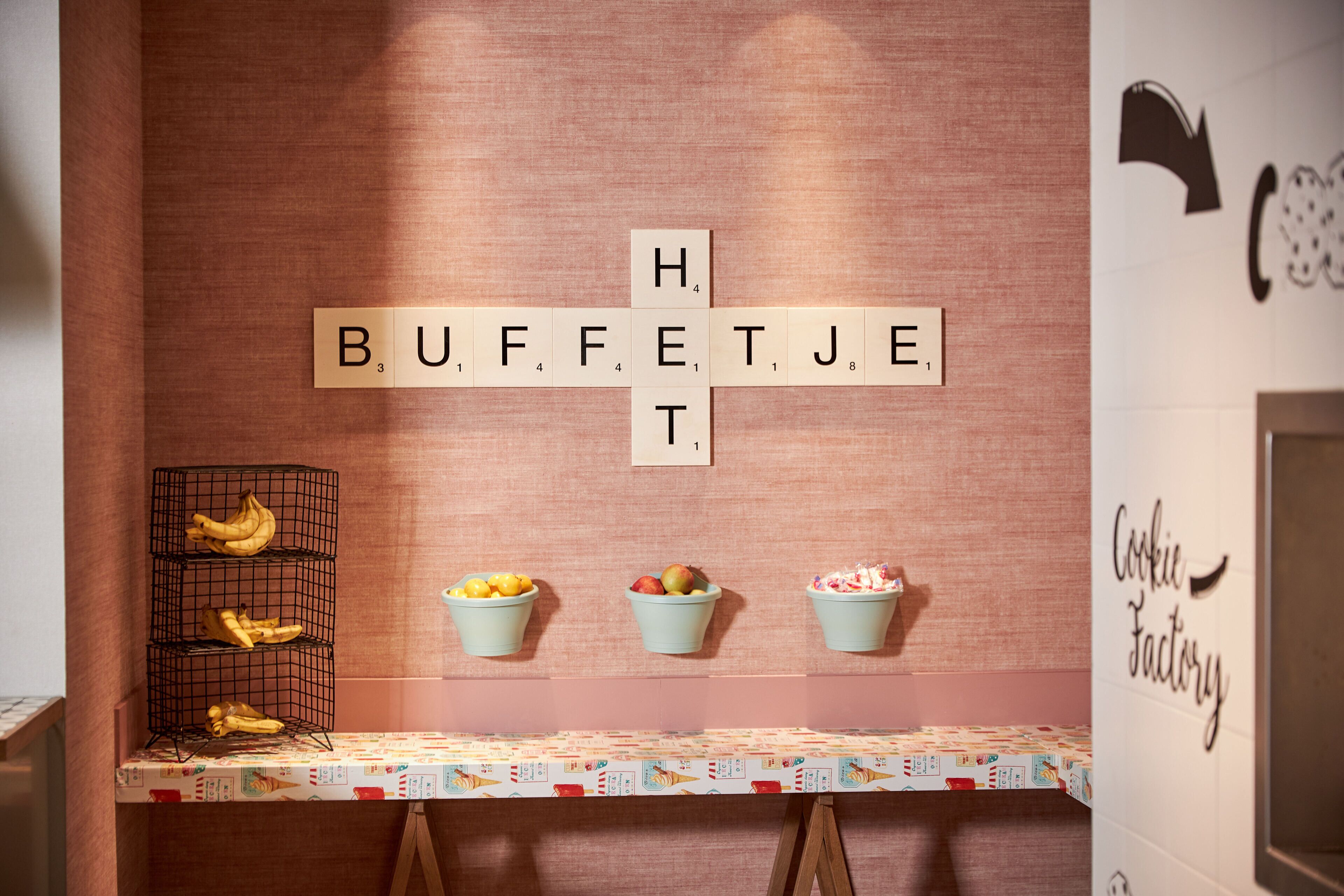 Una zona de cocina con un juego de Scrabble en la pared que deletrea 'BUFFET', una cesta de frutas y un letrero que dice 'Cookie Factory'.