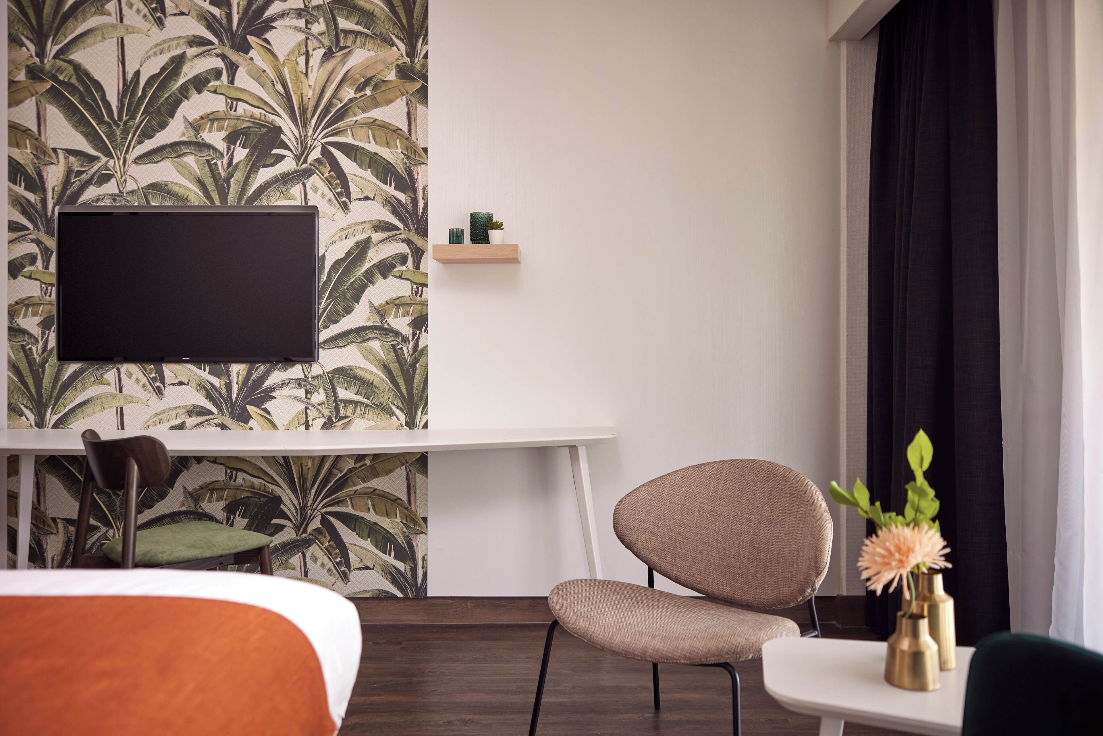 Hotelkamer met een bed, bureau met een flatscreen televisie, stoel, klein tafeltje met een vaas, en een muur met een tropisch bladpatroon.