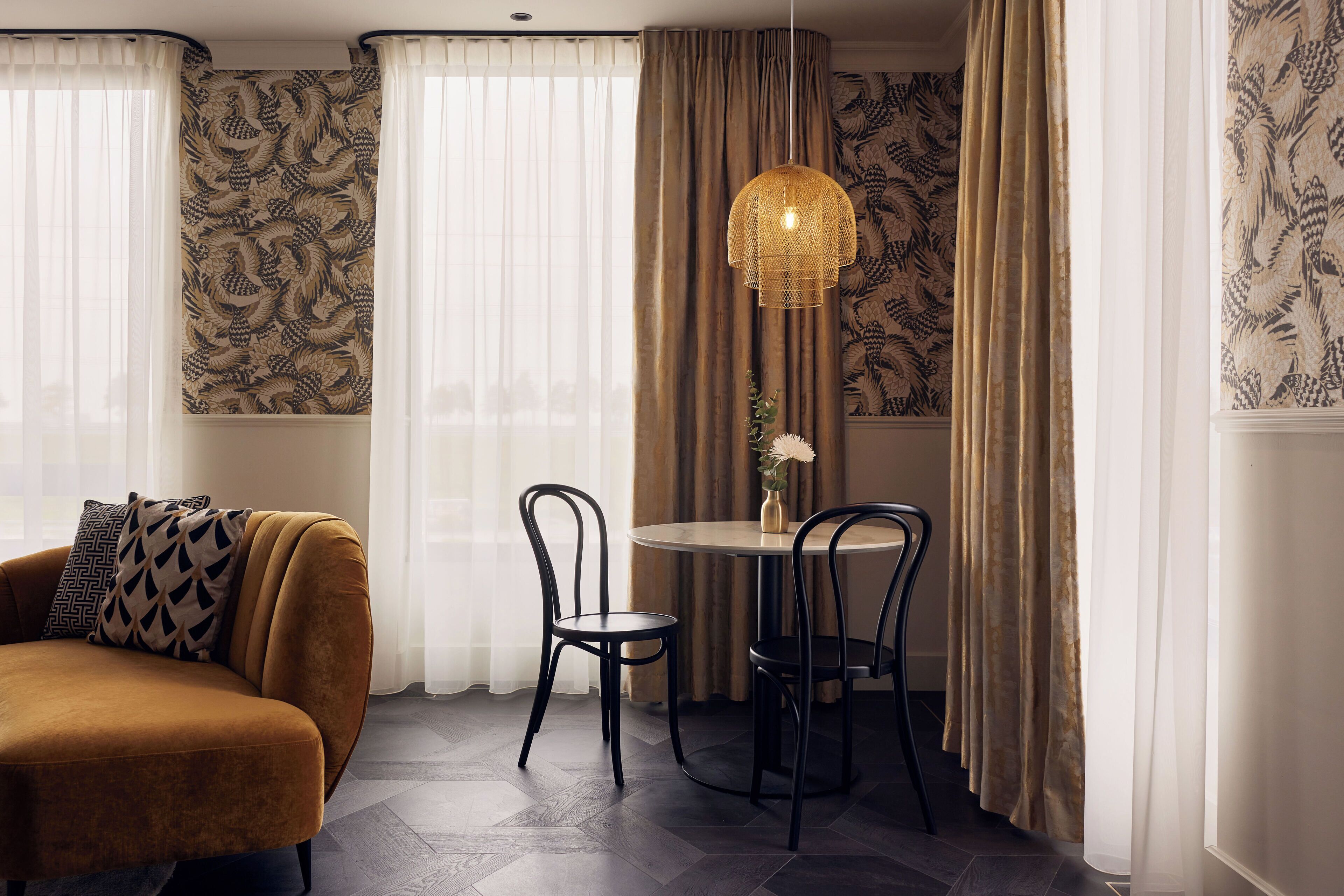 A room with a brown sofa, two black chairs, a round table, a hanging light fixture, and patterned wallpaper.