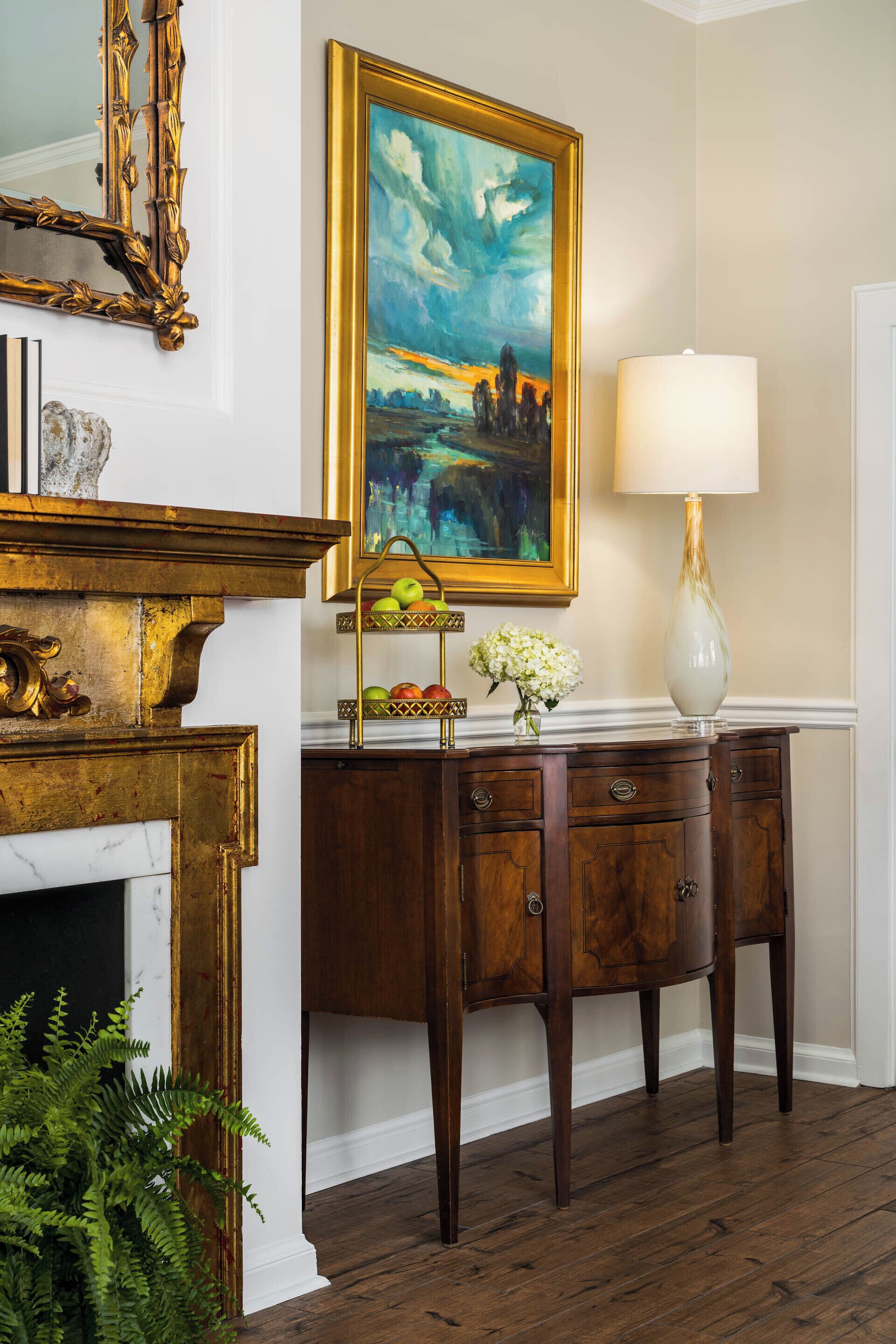 A room with a fireplace, a wooden cabinet with a vase and apples, and a painting on the wall.