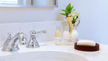 A bathroom sink with two faucets, three bottles of body lotion, and a vase with flowers.