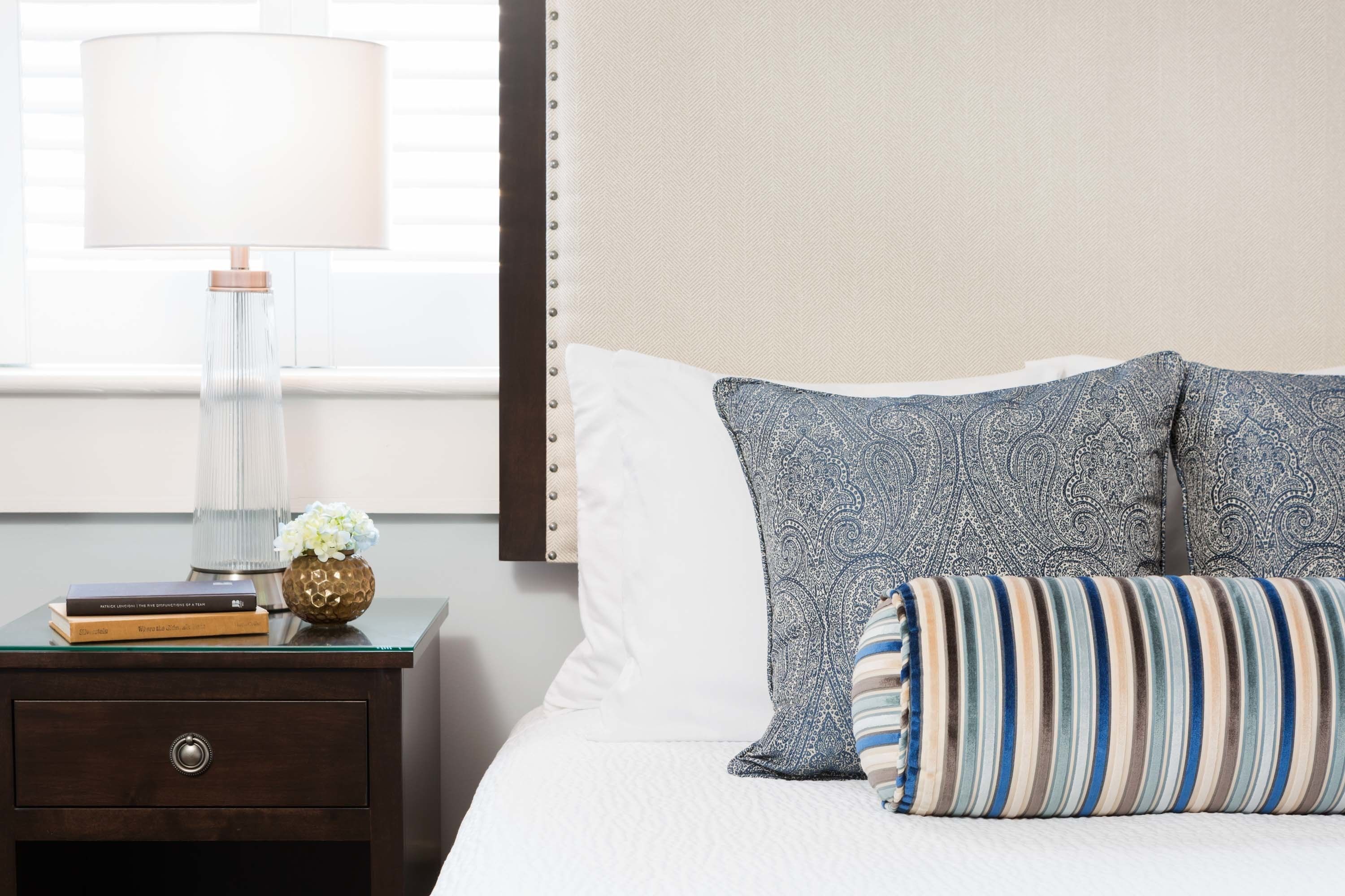 Una cama bien hecha con almohadones estampados, una lámpara de noche, un pequeño florero con flores y una pila de libros sobre una mesita de noche de madera oscura.