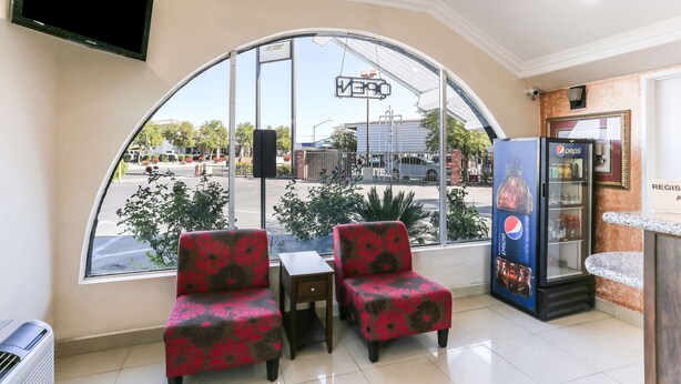 Um saguão de hotel com uma grande janela arqueada, dois poltronas florais vermelhas, uma mesinha, um refrigerador Pepsi e uma televisão de tela plana fixada na parede.