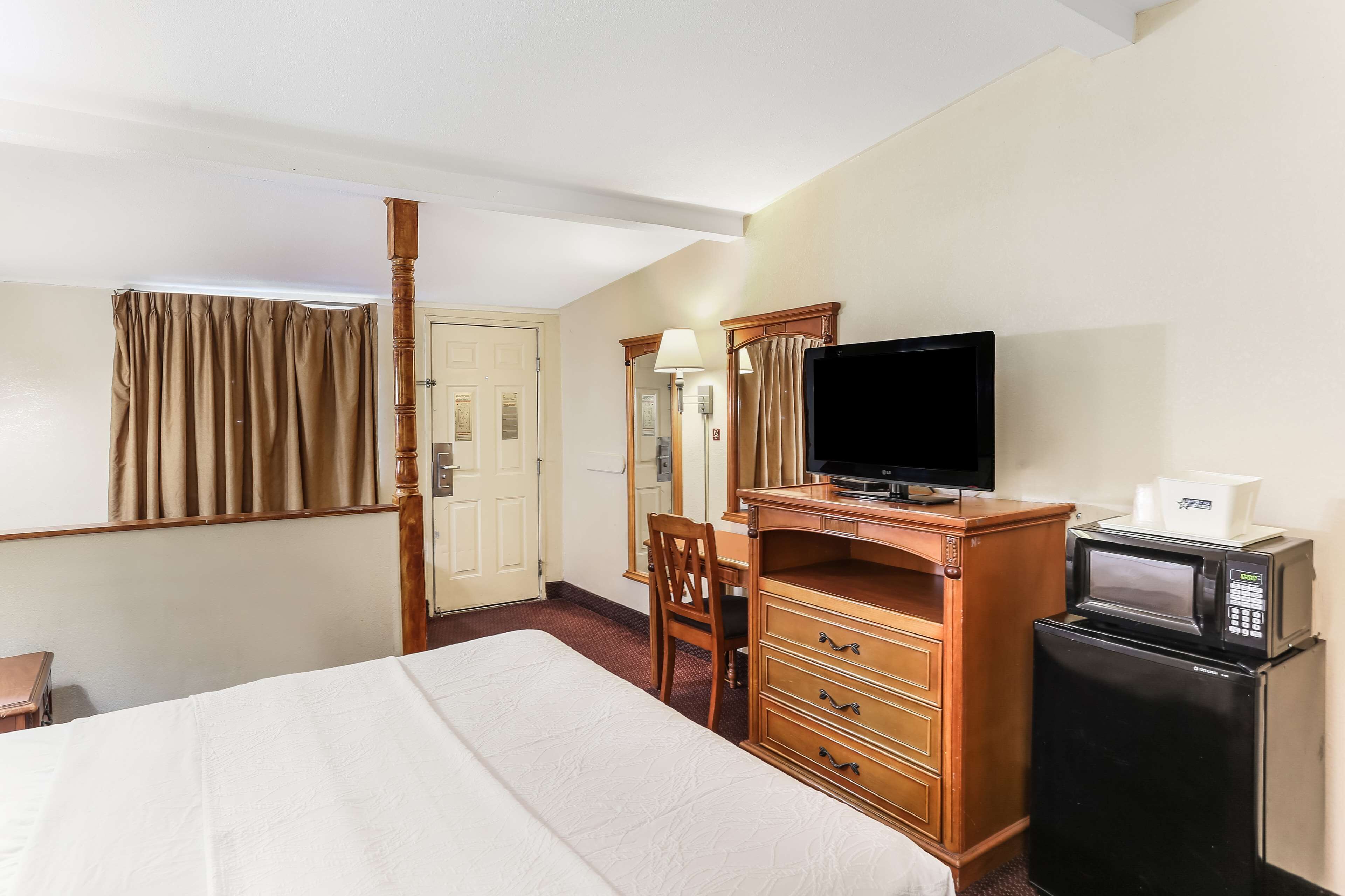 Une chambre d’hôtel comprenant un lit, une télévision, un four à micro-ondes, un réfrigérateur, une commode en bois, une chaise et une porte.