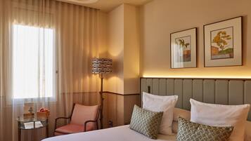 A hotel room with a bed, a chair, a small table with a lamp, and two framed pictures on the wall.