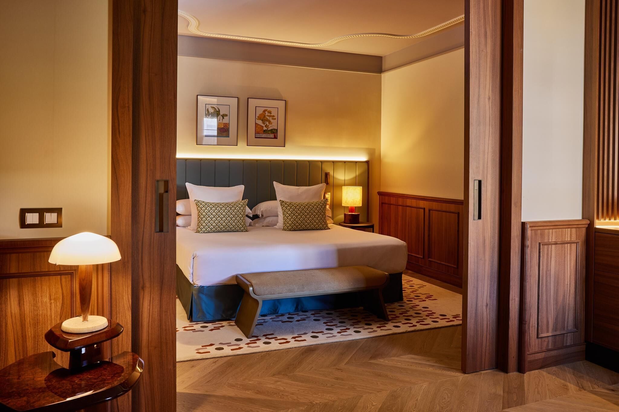 A hotel room with a large bed, a bench, a bedside table with a lamp, and a patterned rug.