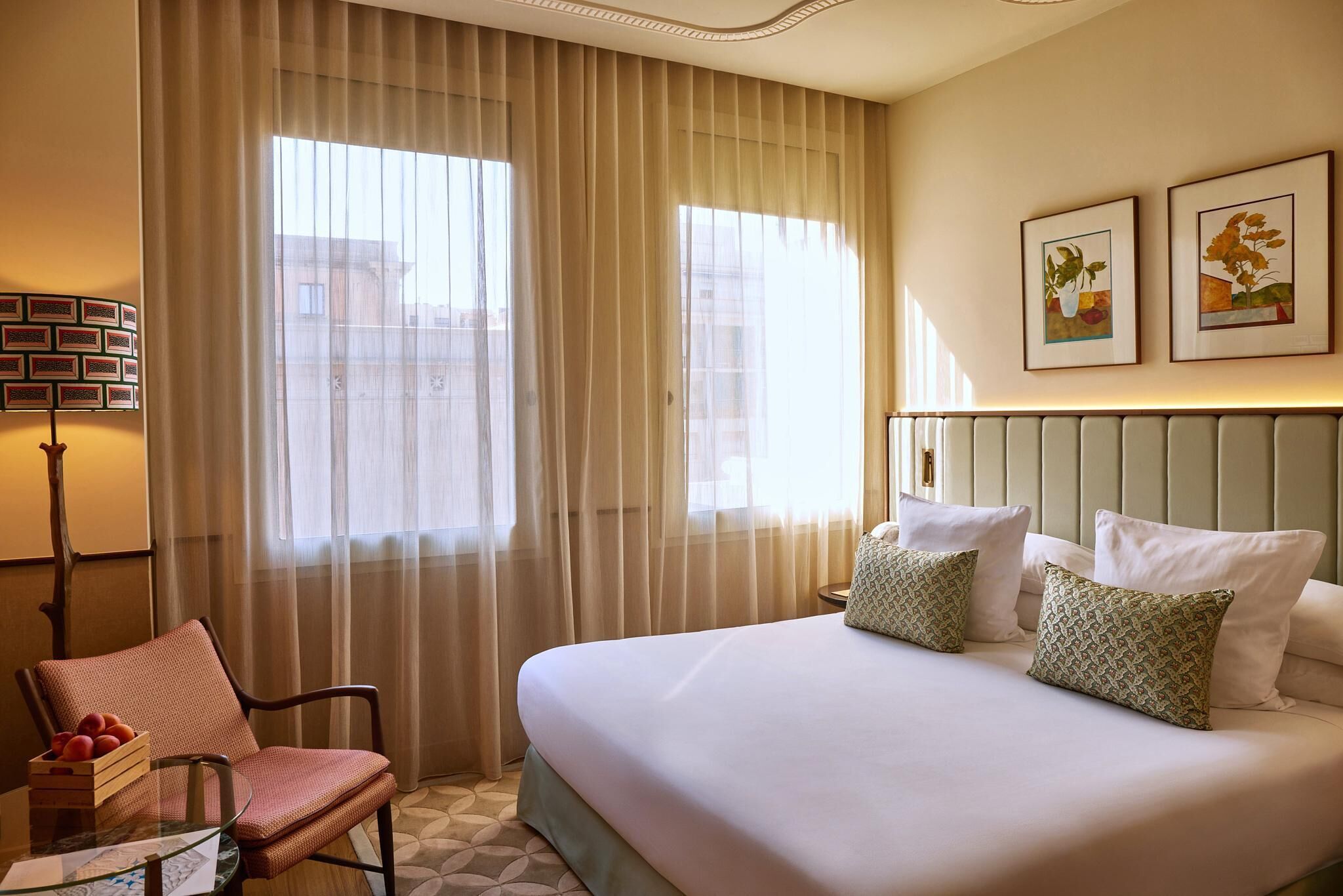 Een hotelkamer met een groot bed, een stoel met een mand fruit, een lamp en twee ingelijste foto's aan de muur.