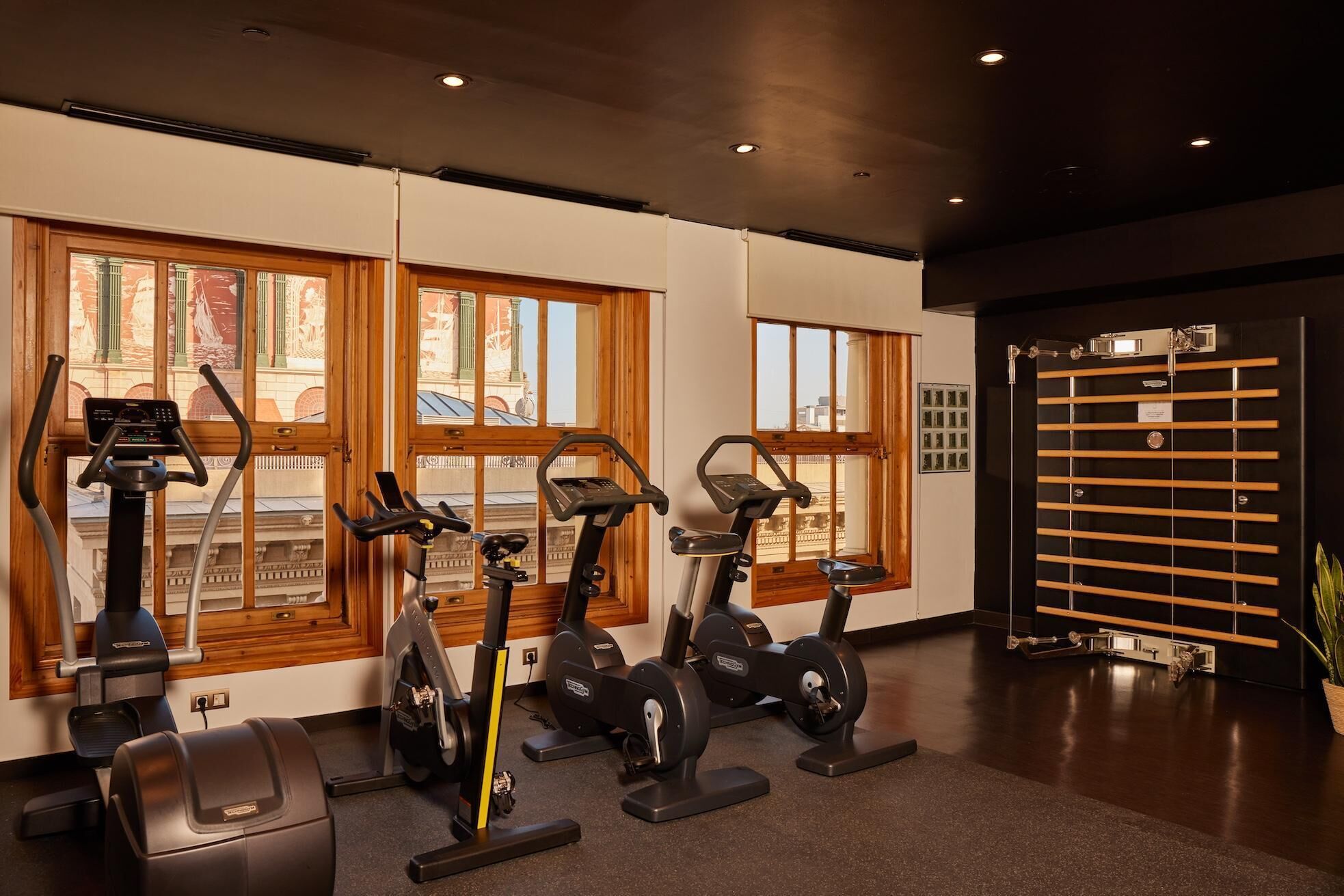 Une salle de sport équipée de quatre vélos d’appartement, de grandes fenêtres donnant sur des immeubles et d’une porte noire avec un portail en métal.