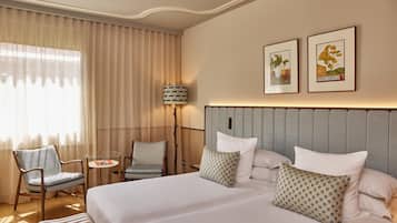 Habitación de hotel con una cama grande, dos sillas, una mesita y una lámpara. Hay dos cuadros enmarcados en la pared.