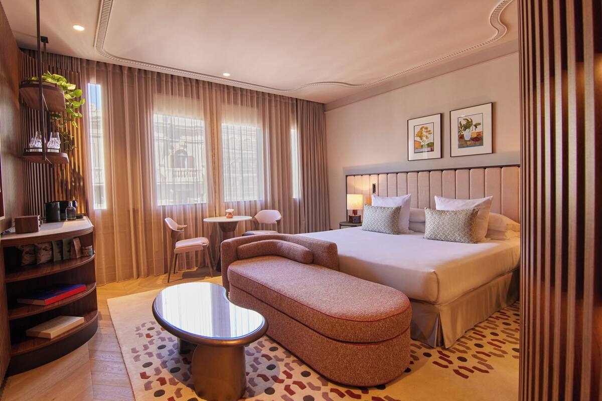 Una habitación de hotel con cama, sofá, una mesita, estantería y ventana con cortinas.