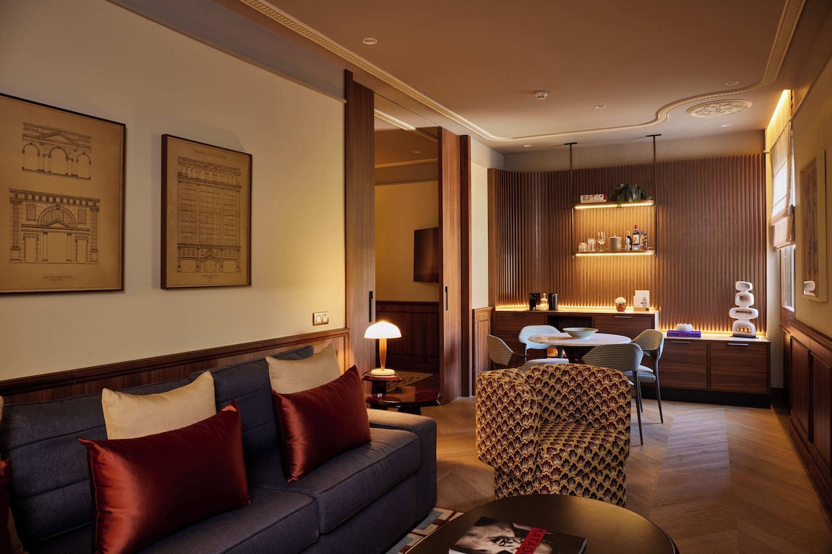 A modern hotel room with a sofa, patterned ottoman, and a dining area with a table and chairs.