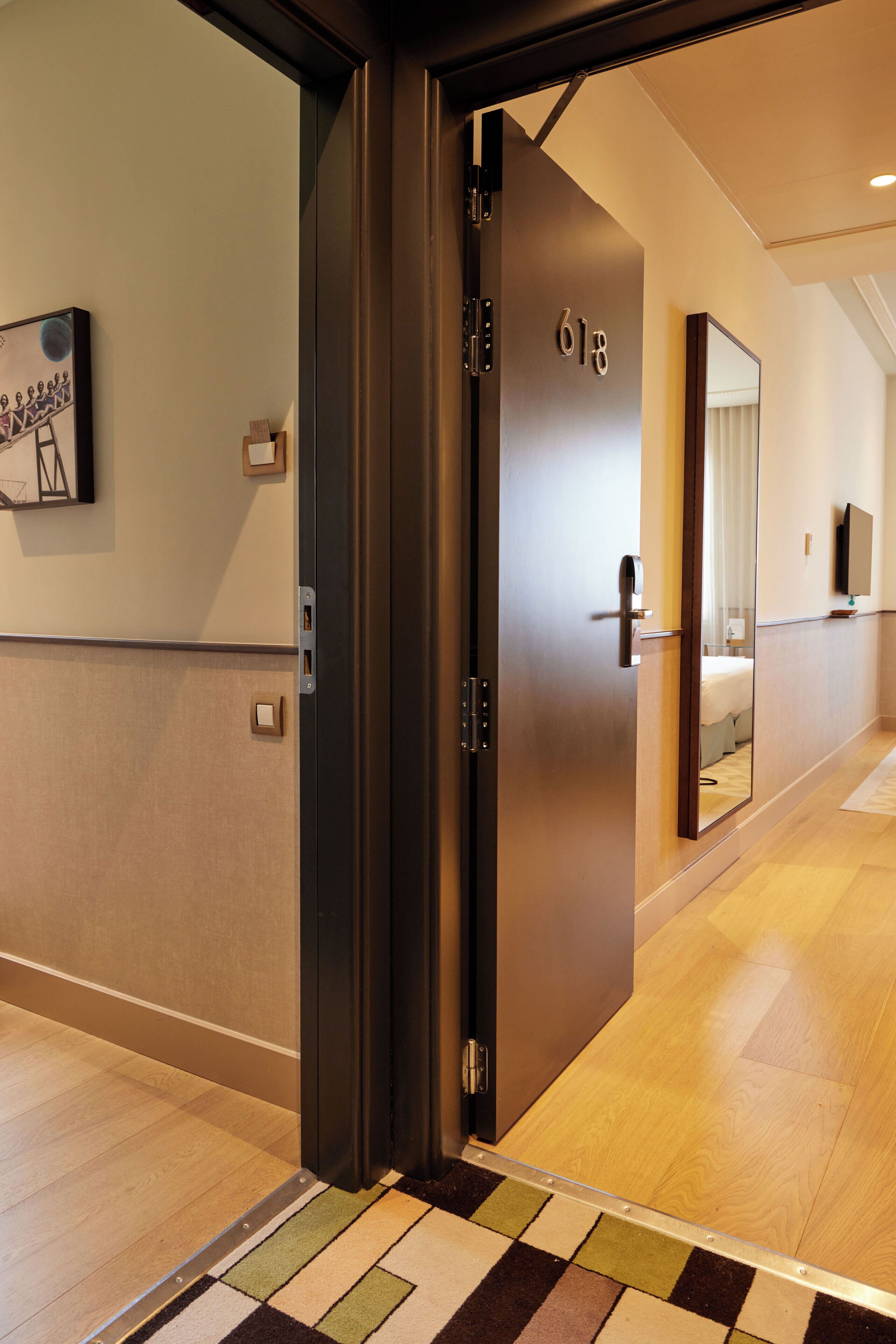 Un pasillo moderno de hotel con suelo de madera, alfombra a cuadros, una puerta con número y un espejo que refleja una cama y un televisor.