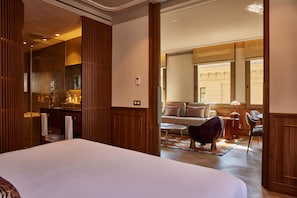 Habitación de hotel con una cama grande, un armario de madera, un baño con ducha de vidrio, una zona de estar con sofá y sillones, y vistas del exterior del edificio.