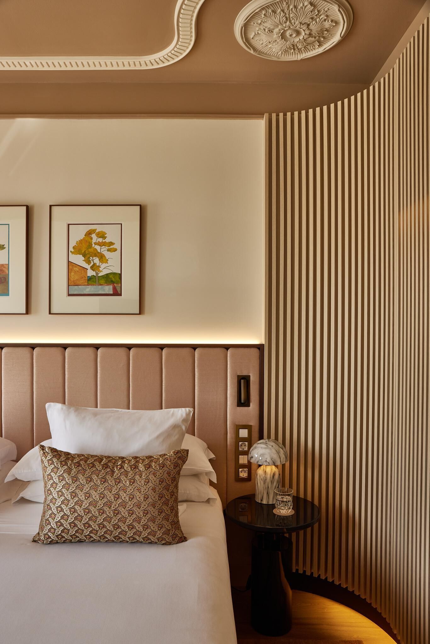Una habitación de hotel con una cama grande, un cabecero, una mesita de noche con una lámpara y dos cuadros enmarcados en la pared.