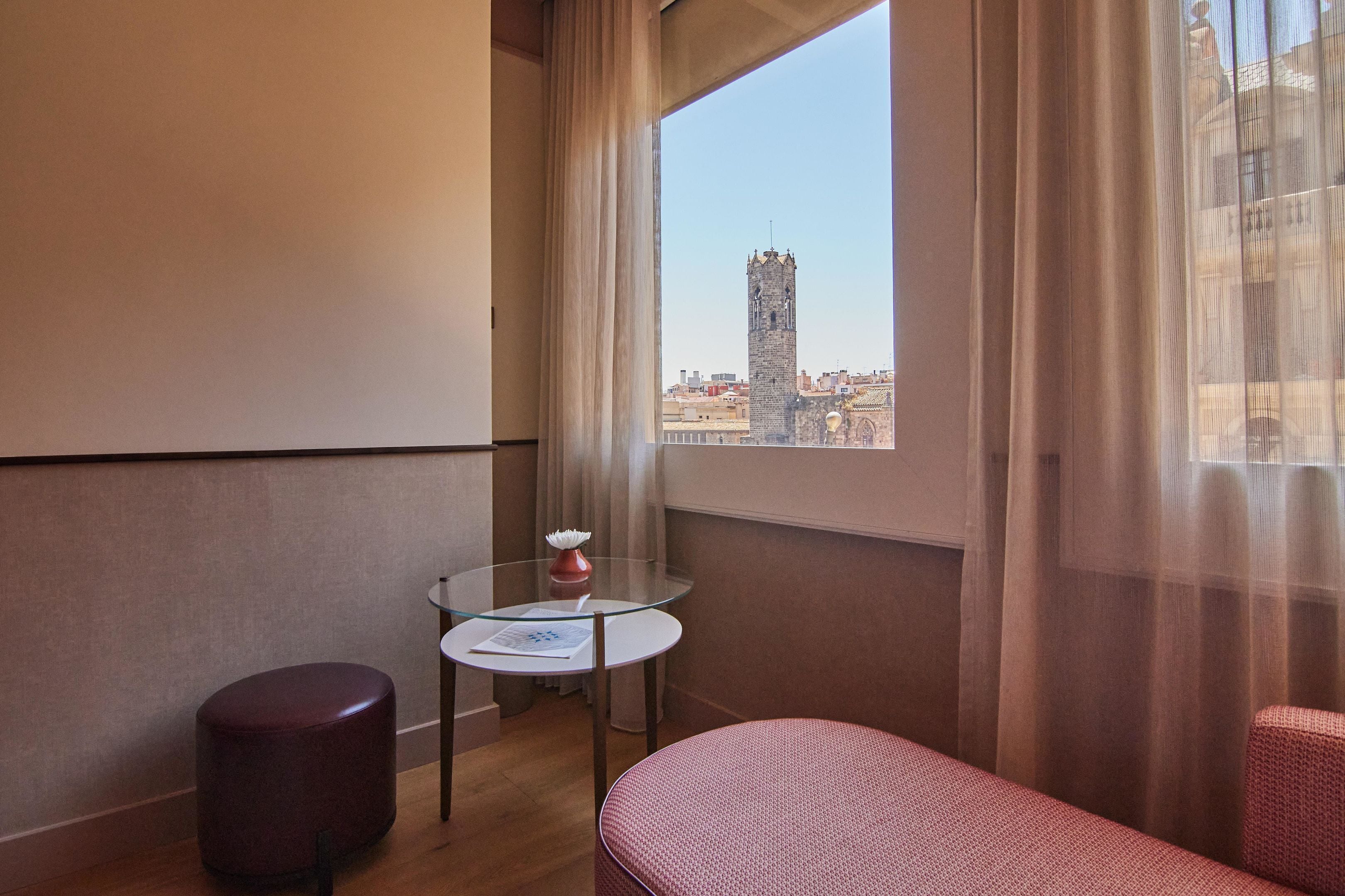 Une chambre d’hôtel avec une table en verre, un pouf rouge et une vue sur une haute tour, visible à travers de fines rideaux.