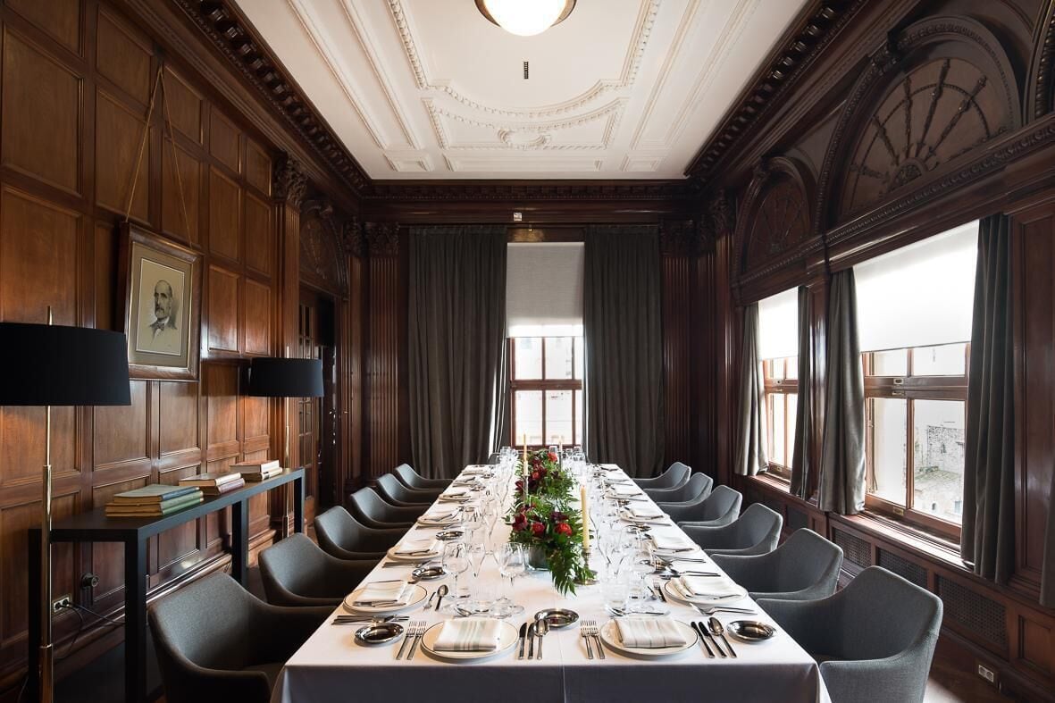 Una larga mesa de comedor preparada para una comida formal en una habitación con paneles de madera y ventanas amplias.