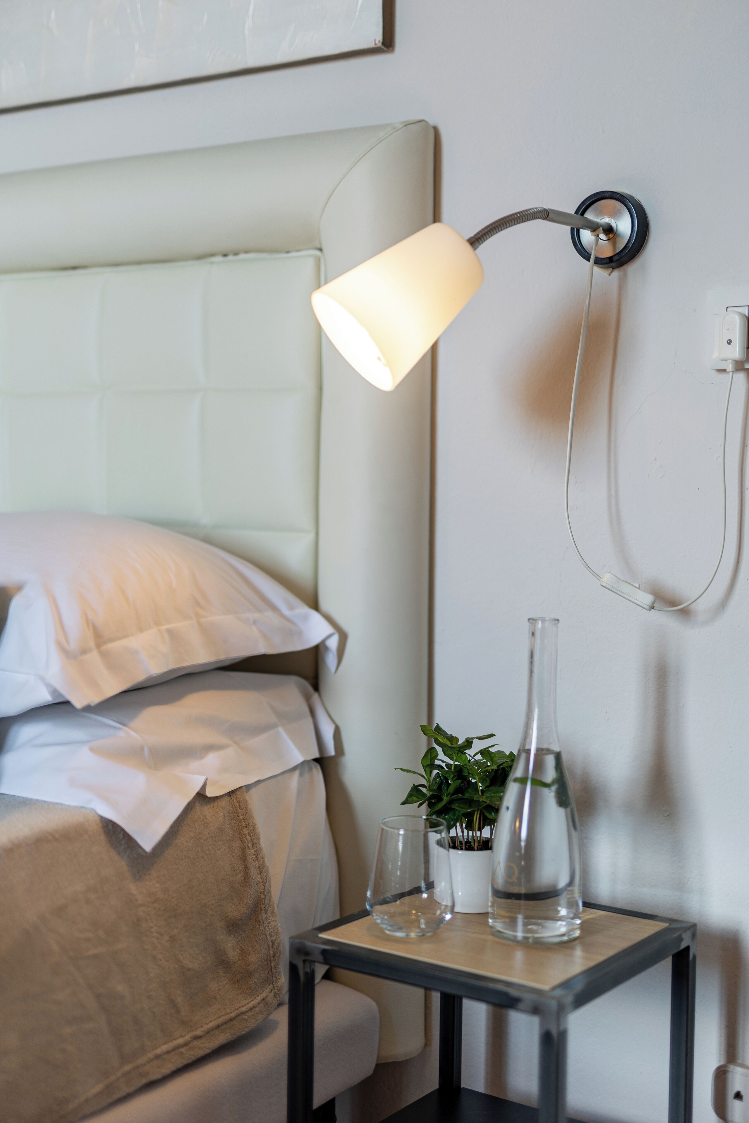 En pænt redt seng med hvide sengetæpper, et natbord med en klar glasflaske og en lille urtepotte, samt en vægmonteret lampe.