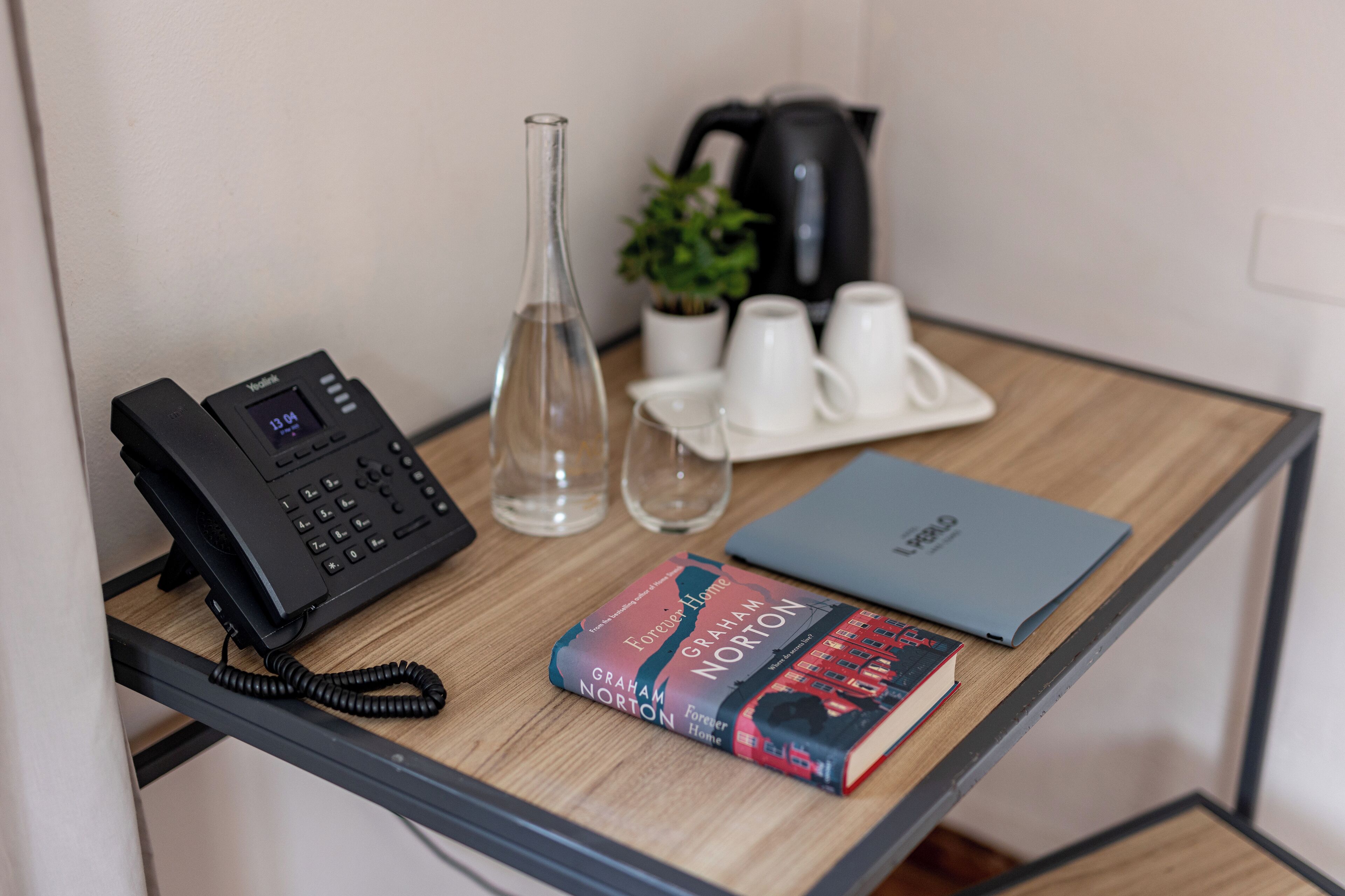 Een houten bureau met een zwarte telefoon, een glazen vaas, een potplant, een dienblad met witte bekers, een boek getiteld 'Forever Home' van Graham Norton, en een blauw notitieboek.