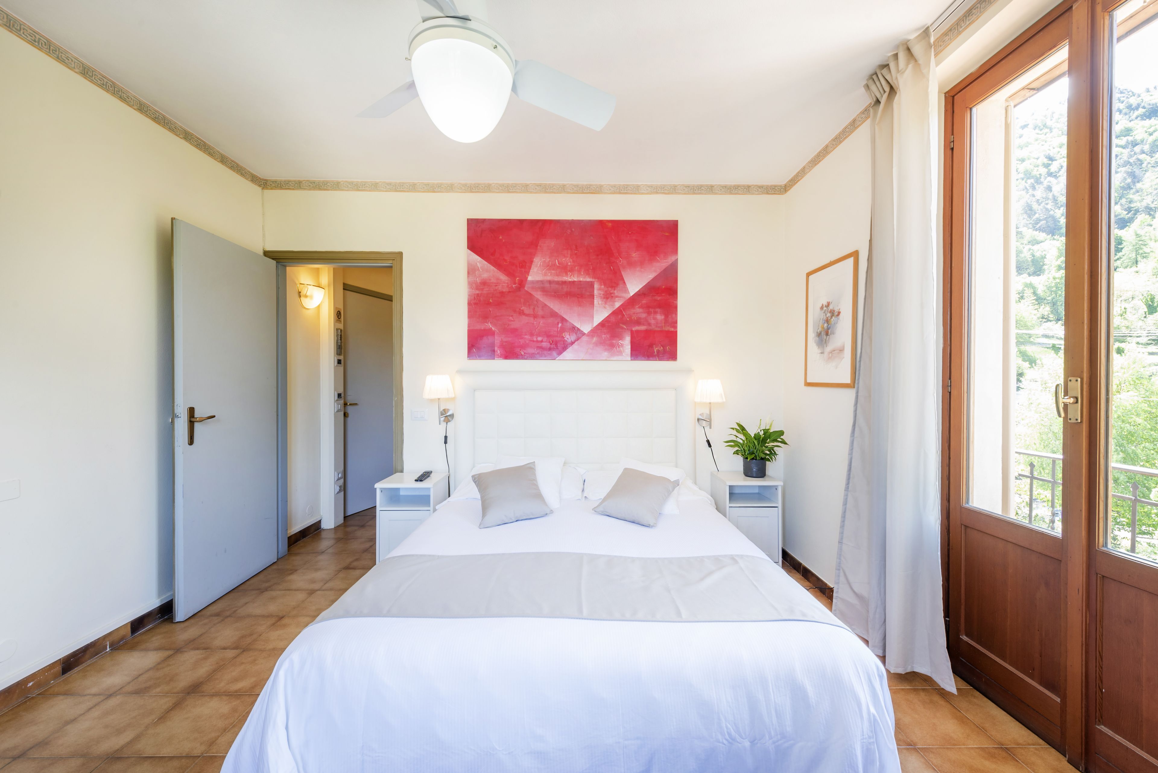 A bedroom with a bed, a ceiling fan, a red abstract painting, a potted plant, and a view of greenery through a window.