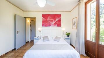 A bedroom with a bed, a ceiling fan, a red abstract painting, a potted plant, and a view of greenery through a window.