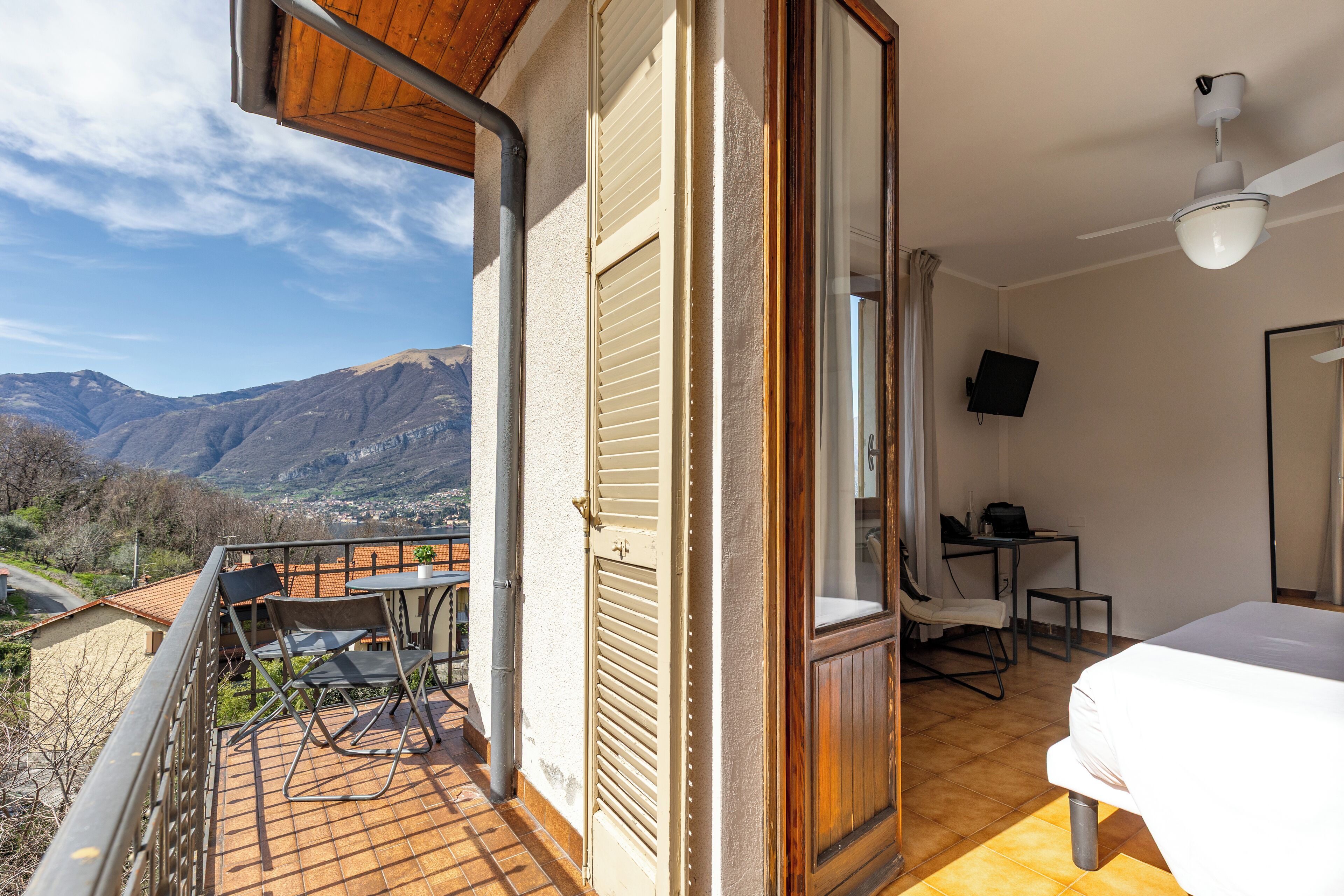 Una habitación con cama, escritorio y silla. Tiene un balcón con vistas a montañas.